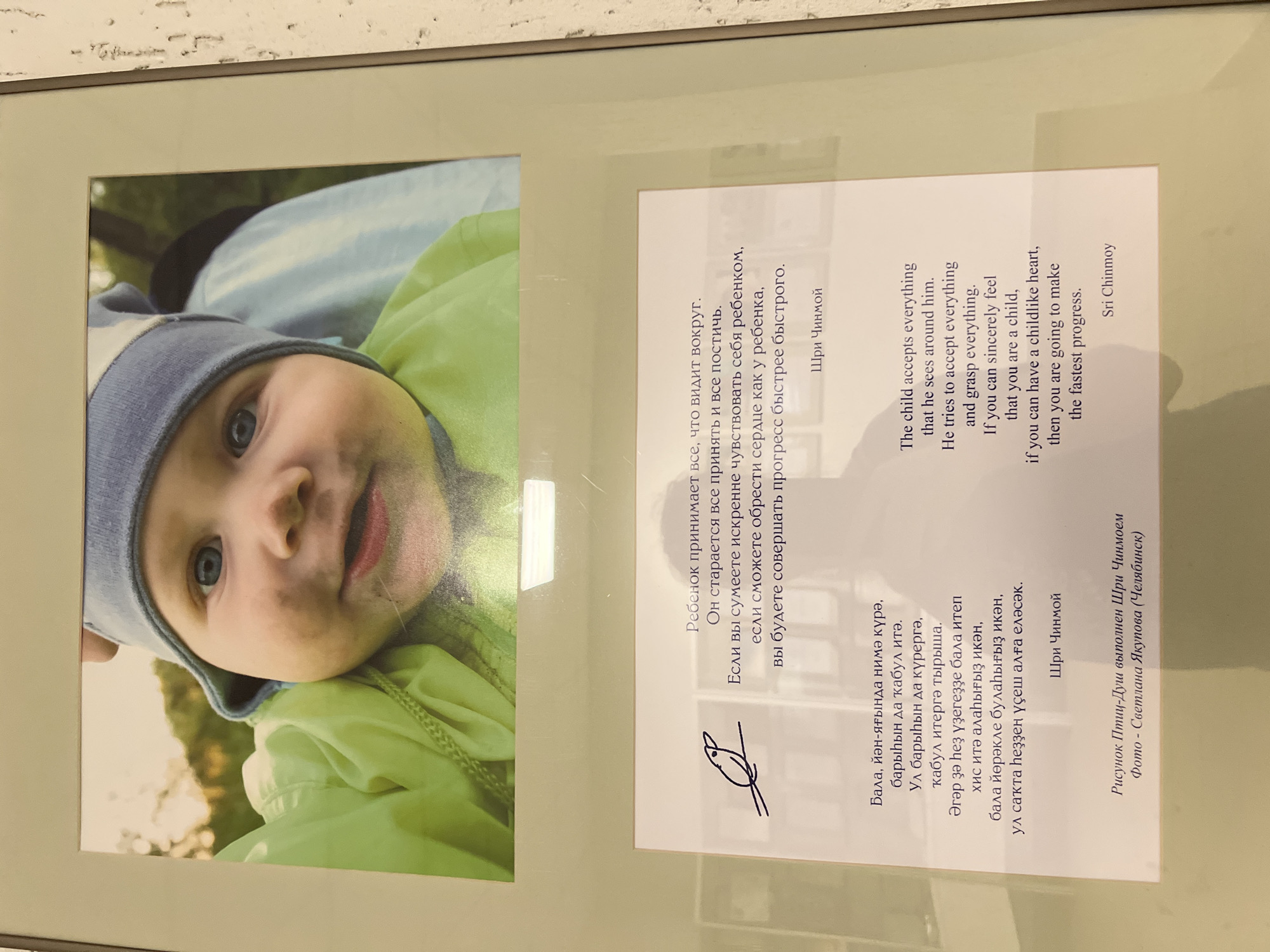 The image is a framed picture hanging on a textured white wall. The picture features a close-up photograph of a young child with blue eyes, wearing a blue hat and green jacket. To the right of the child's photo is text in Russian and English. The text is a quote by Sri Chinmoy about a child's perspective and the ability to make progress by having a childlike heart. Below the text is a drawing resembling a bird and more text identifying the drawing as being by Sri Chinmoy and the photograph as being taken by Svetlana Yakupova from Chelyabinsk. The photo was taken in the city of Sanatoriya Yumatovo Imeni 15-Letiya Bassr, Russia.