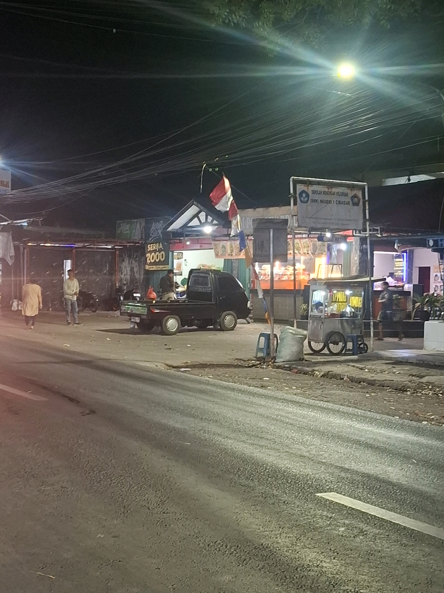 The image depicts a street scene at night in Karangtengah, Indonesia, possibly near a school given the sign that reads "SEKOLAH MENENGAH KEJURUAN (SMK) NEGERI 1 CIBADAK". The street is paved with asphalt, and a white line is visible marking a lane. On the right side of the street, there is a food cart with yellow signage and two blue stools. Behind the food cart, a person in dark clothing is standing. Further back, a building with illuminated windows suggests a shop or eatery. On the left side of the street, a black pickup truck is parked. Two individuals are standing near the truck, one in a light-colored shirt and pants, and another further back in a beige outfit. A large white sack is positioned next to the food cart. Overhead, a network of power lines crisscrosses the dark sky, illuminated by a bright street lamp casting a green-tinged glow. A red and white Indonesian flag is visible on a pole above a structure. The general environment suggests a commercial or semi-commercial area in a town or city.