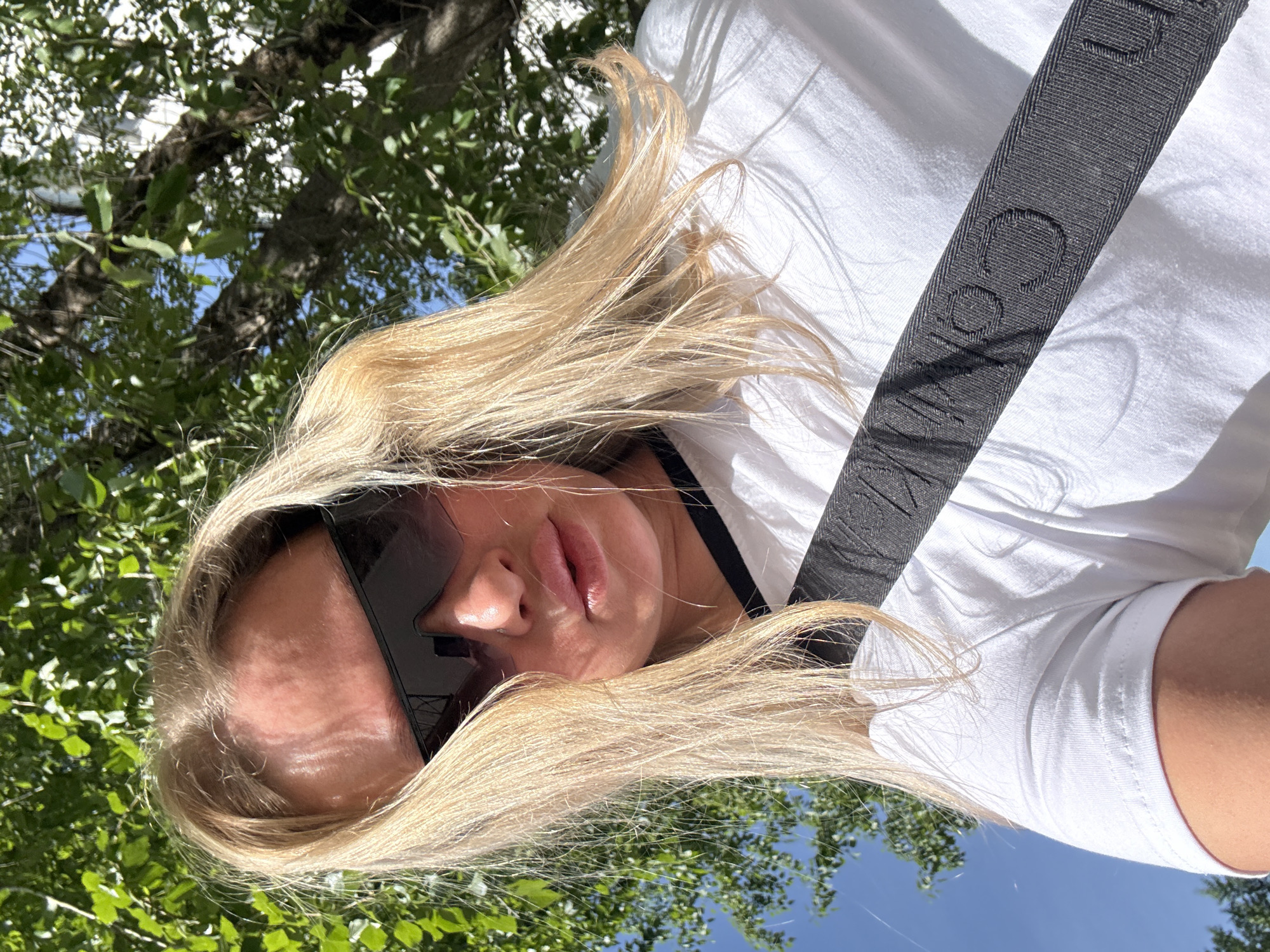 A woman with blonde hair, wearing black sunglasses, a white t-shirt, and a black seatbelt with white writing, is looking up at the camera from a low angle. The background includes trees and blue sky. The location cannot be confirmed without further information.