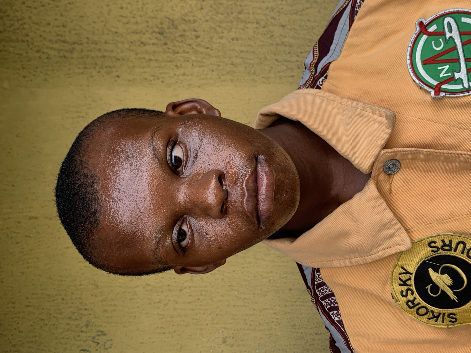 A portrait of a Black man is positioned against a textured yellow background. The man has a close-cropped haircut and his skin appears to have a glossy sheen. He is wearing a tan collared shirt or jacket. There are two circular patches on the jacket. The first patch, on the right shoulder, has the words "SIKORSKY B-SKY HOURS" written around the border of a black center that features a flying helicopter with outstretched wings. The second patch on the upper right chest, contains the letters "N," "V," "C," and "Z" in white with green background. The man's expression is neutral. The scene provides no clues about a specific location in Ikorodu, Nigeria.