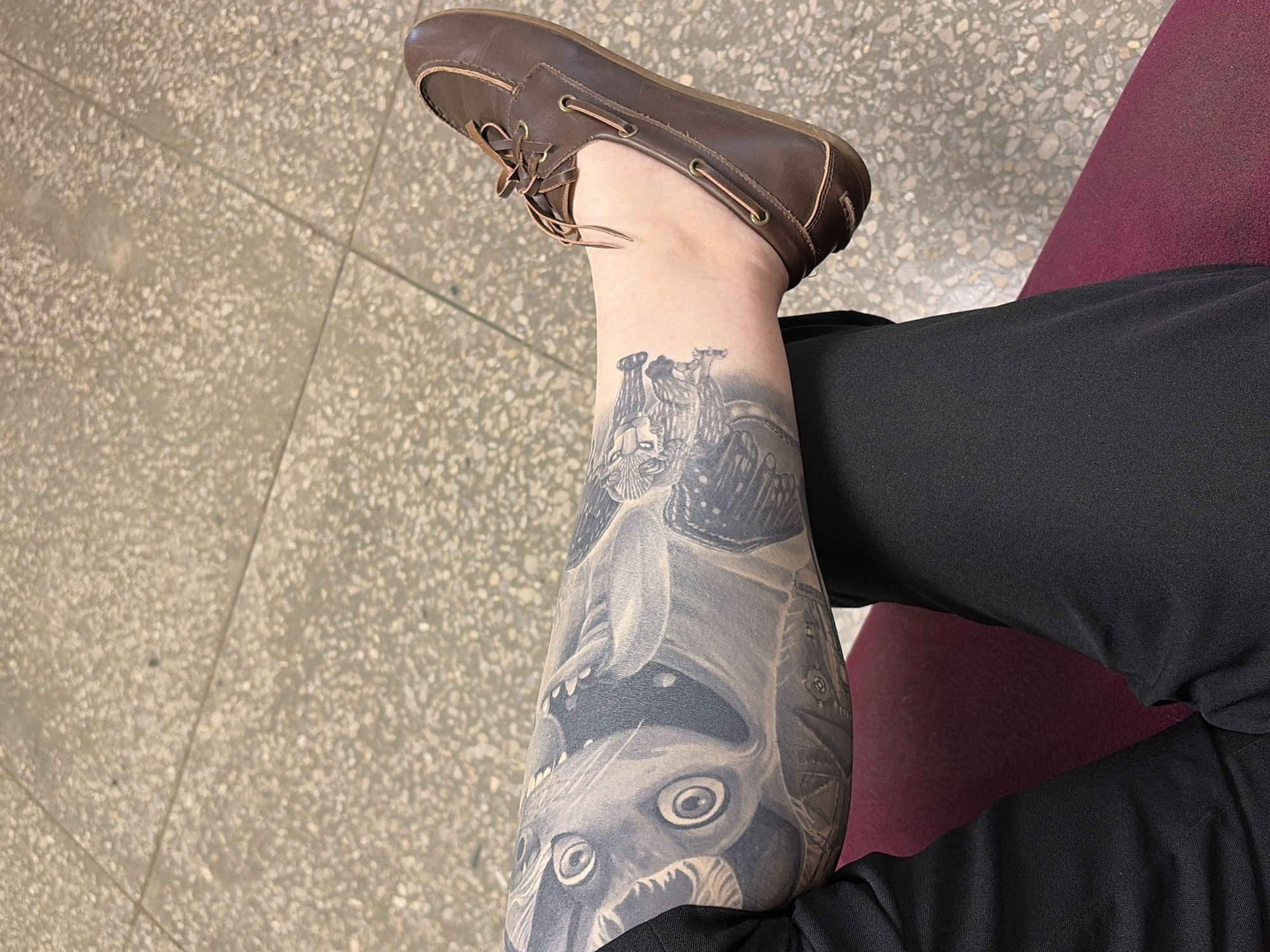 A close-up shot shows a person's lower leg and foot resting on what appears to be a cushioned surface. The person is wearing dark trousers and a brown leather boat shoe. A detailed tattoo covers a significant portion of the lower leg. The floor in the background is tiled and appears to be in an indoor location. Based on the tattoo's imagery (a city scene resembling a Russian architectural style), and the request to connect it to Chaikovsky, Russia, it can be surmised that the person may be in that city. However, the image itself provides no definitive proof of the location, and the statement is based solely on circumstantial information.