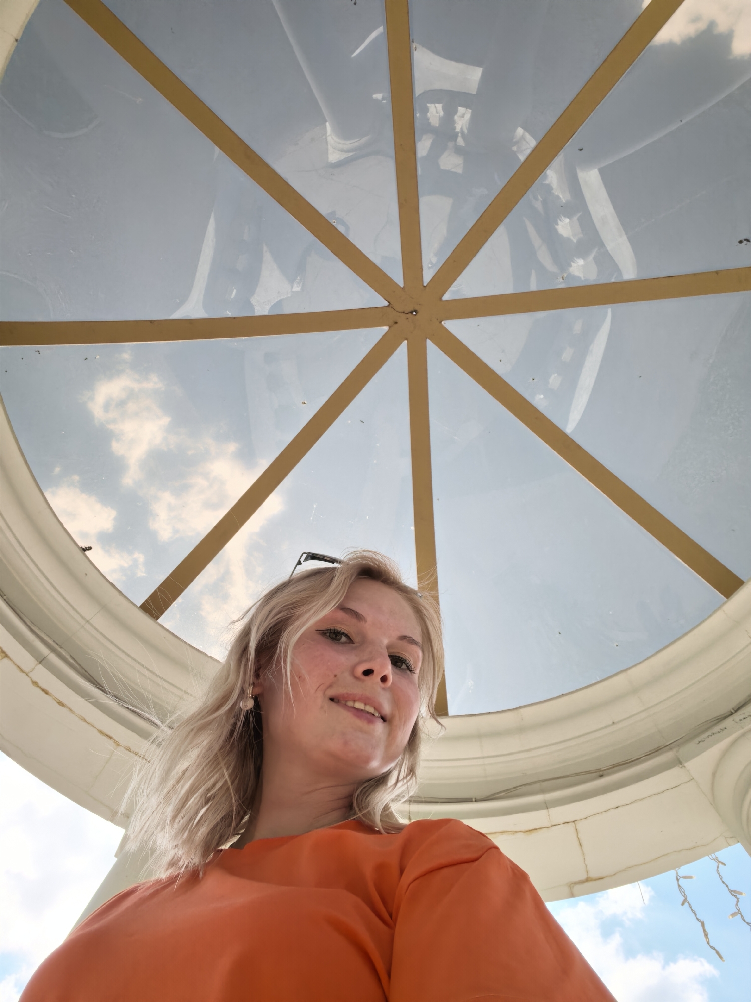 A young woman with blonde hair is captured in a low-angle selfie, looking upwards at a glass dome with golden metal supports. She is wearing an orange t-shirt and has a pair of sunglasses perched on her head. The dome's structure is visible, with light reflecting off its surface and the sky visible through it, showing scattered clouds. The surrounding architectural element appears to be a white, curved structure with some visible wires or string lights hanging down. The setting is likely outdoors, under a covered structure with a domed roof. There is no specific information linking this image to the city of Sterlitamak, Russia.