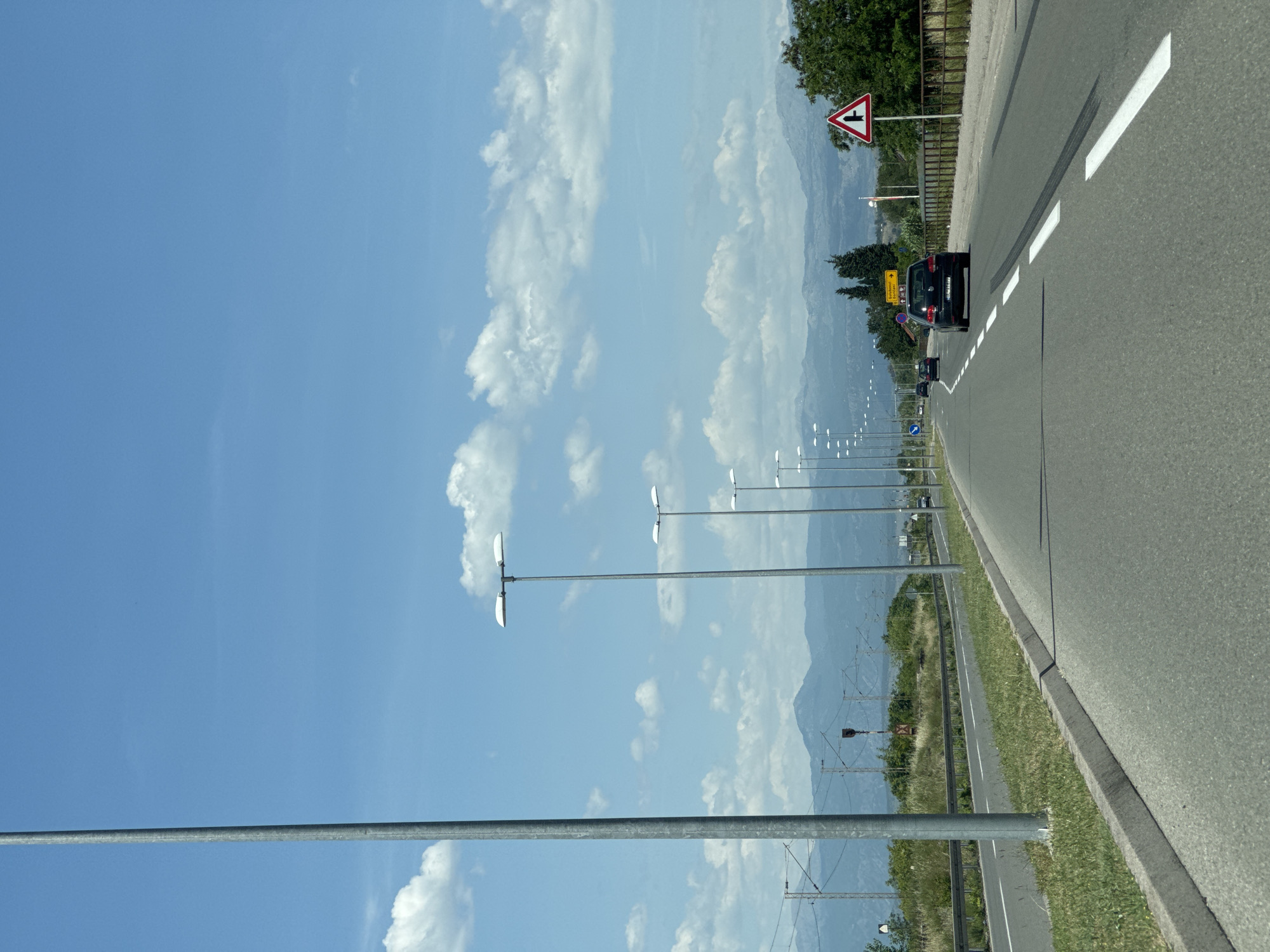 The image depicts a road running through Goričani, Montenegro, with mountains visible in the background. Several lampposts line the road. A car is present on the road, along with a road sign indicating a junction. The sky is blue with scattered clouds. Overhead power lines and supporting structures are also visible. The road has painted lane markings.