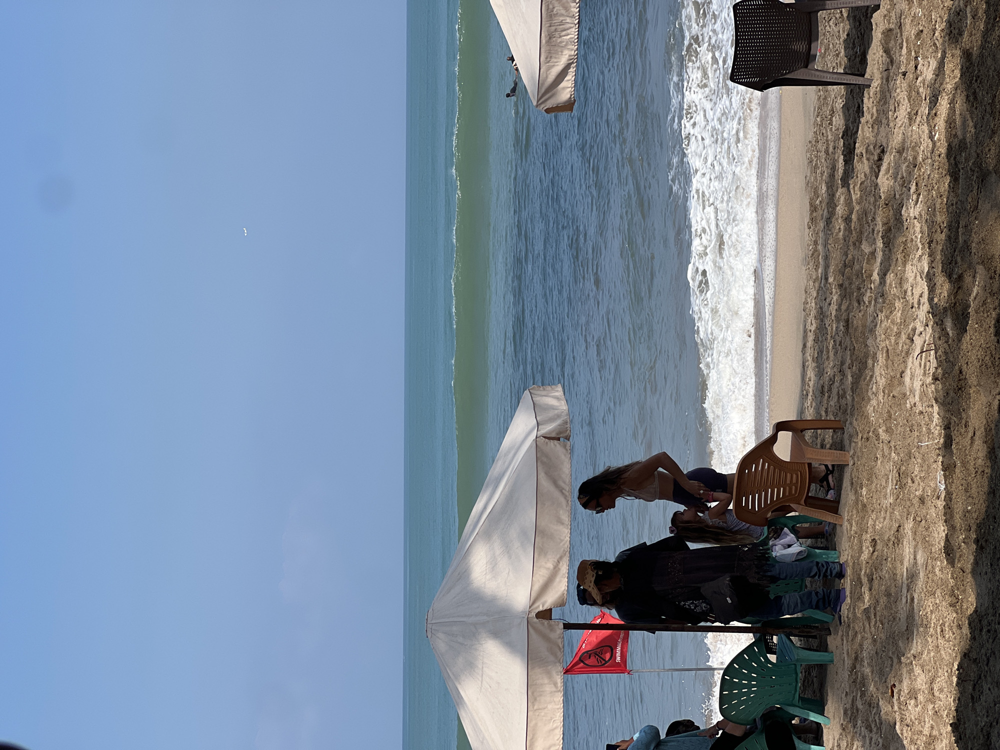 The image depicts a beach scene in Kuta, Indonesia, likely during the daytime, as indicated by the bright sunlight and clear blue sky. The foreground is dominated by sand, with several people gathered near beach umbrellas and chairs.
The subjects include at least three individuals, two women and one person whose gender is less clear, positioned under a large white beach umbrella. One woman, wearing a white top and dark bottoms, appears to be leaning down or interacting with someone or something near the ground. Another woman, wearing a white bikini top and dark bottoms, is looking down. A third person, wearing a dark top and jeans, is also under the umbrella. Several beach chairs, including a brown plastic one and green plastic ones, are scattered around the area. A red flag with white text is visible, attached to a pole.
The activity taking place seems to be a leisurely day at the beach. People are relaxing, sitting, and possibly socializing. The ocean is in the background, with waves gently rolling onto the shore. The water appears a clear blue-green color, and the waves are creating white foam as they break. The overall atmosphere appears relaxed and casual, consistent with a beach resort location like Kuta.
Notable details include the presence of beach umbrellas, chairs, and the ocean, which are characteristic of a beach setting. The sand appears somewhat wet near the waterline. The lighting suggests it's a sunny day, possibly mid-morning or afternoon. While the exact emotions are hard to discern, the relaxed postures suggest a comfortable and pleasant experience. The text on the red flag is partially visible and appears to be a warning or informational sign, though it's not fully legible.