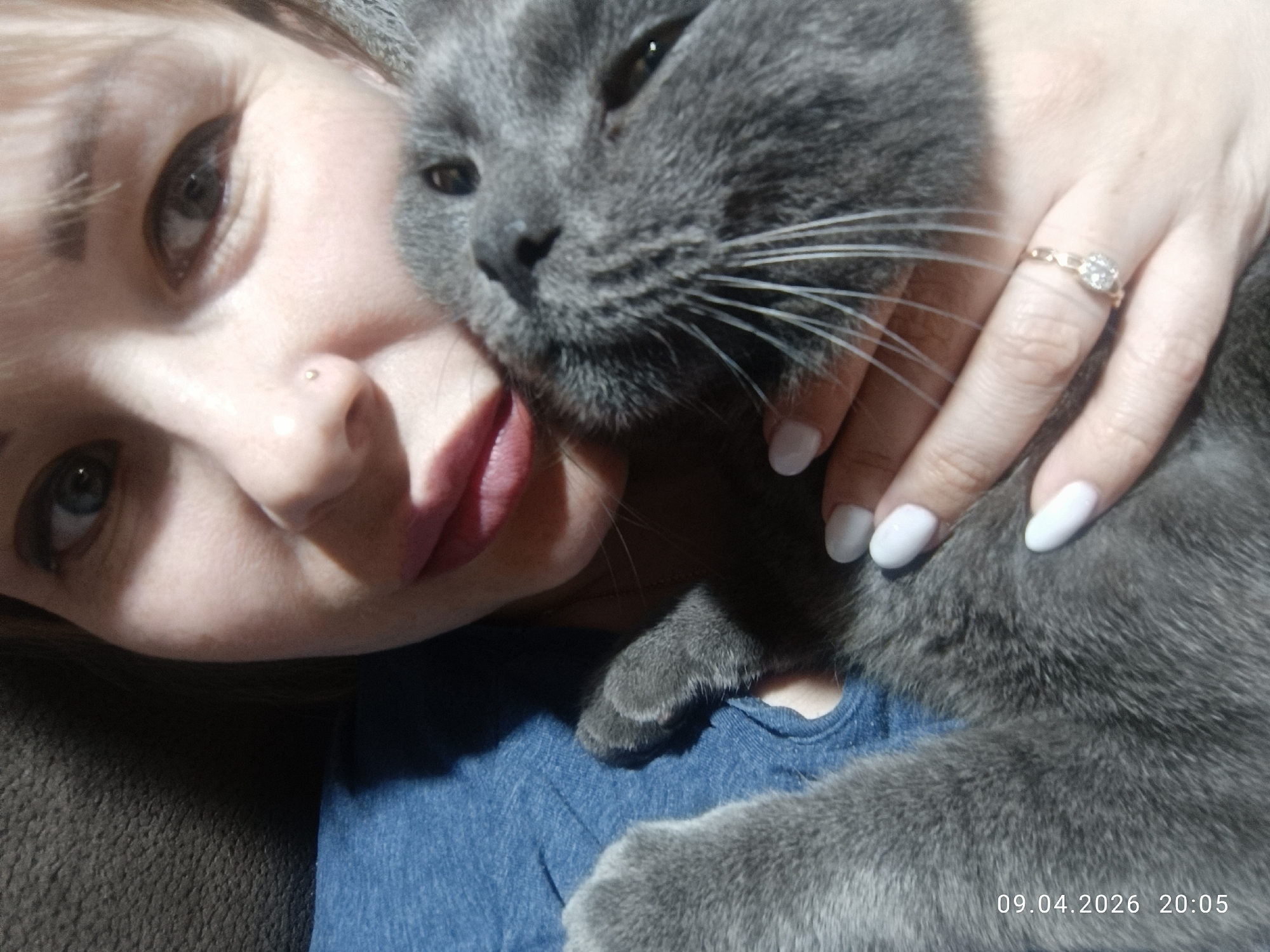 A woman with light-blonde hair and a nose piercing is cradling a gray cat in her arms. She is wearing a blue denim shirt. Her left hand is visible, and she has a ring on her ring finger. The date and time, "09.04.2026 20:05," are printed on the lower right corner of the image. The image was taken in the city of Chaikovsky, Russia.