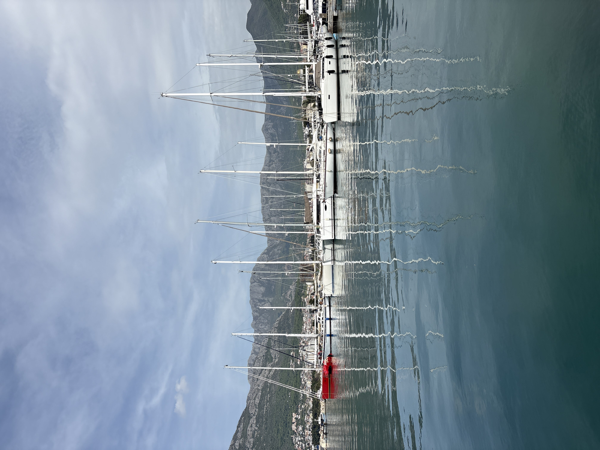 The image, correctly oriented, presents a tranquil harbor scene in Bar, Montenegro, featuring a marina filled with numerous sailboats backed by mountains and a cloudy sky.
In the foreground, the calm, expansive water of the marina takes center stage. Its surface, ranging from light blue-grey to a deeper teal, beautifully reflects the masts and hulls of the docked boats, creating abstract, wavy patterns that ripple across the water.
A prominent line of sleek sailboats, primarily white, are moored closely together along the pier. Their tall, slender masts rise vertically into the sky, creating a sense of ordered rhythm. One boat, distinctly colored in bright red, stands out among the white vessels in the lower-left section of the image, adding a vibrant focal point. No specific activity or people are visible, indicating a peaceful moment at the dock.
Behind the marina, the midground reveals a coastal town nestled at the foot of a grand mountain range. Buildings of various sizes are visible across the lower slopes, illustrating the blend of urban development with the natural environment. The mountains themselves appear rugged and majestic, providing a dramatic, verdant backdrop to the harbor.
The sky above is a soft, light blue, dappled with thin, wispy white clouds. The overall lighting suggests it is daytime, likely late morning or early afternoon, with a gentle, diffused light. There is no visible text in the image. The scene conveys a sense of serenity and the picturesque beauty of a Mediterranean or Adriatic port.