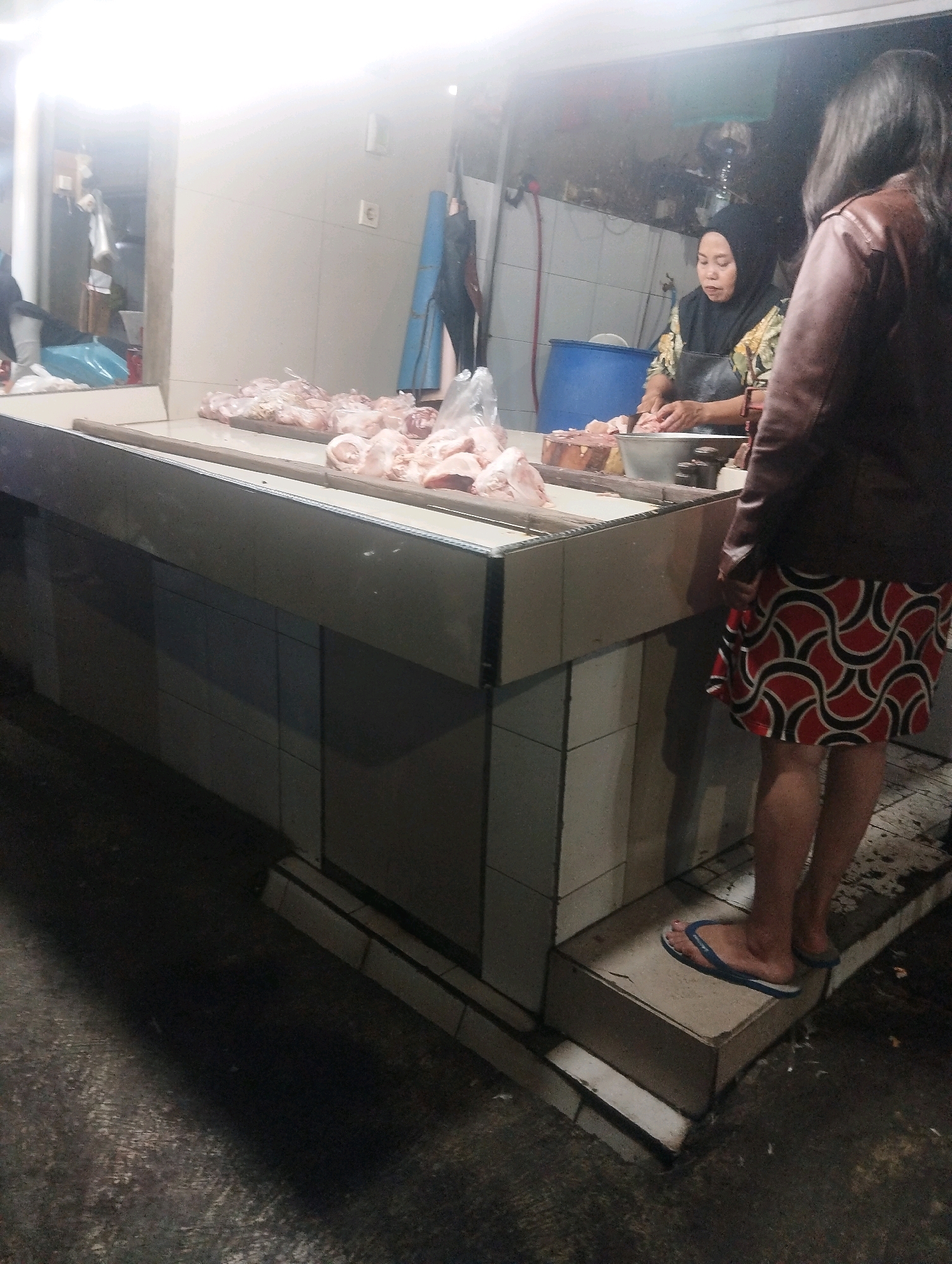 The image depicts an outdoor market stall in Malang, Indonesia, likely selling meat. Two women are present. One woman, wearing a headscarf and apron, is actively working behind the counter, seemingly preparing or cutting meat. She is positioned in front of a large blue barrel. The second woman, facing away from the camera, is standing to the right of the stall, wearing a patterned skirt and a jacket. The stall itself is a long, rectangular counter displaying various cuts of meat. The environment appears to be a covered area, with bright overhead lighting illuminating the scene and casting shadows on the dark, textured ground. The overall impression is of a bustling, functional marketplace.