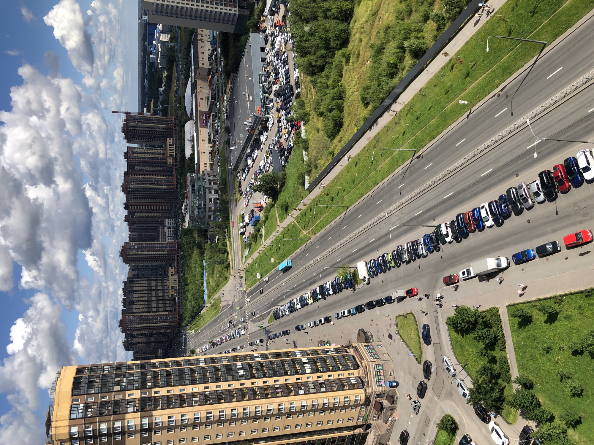 An aerial view captures a sunlit urban environment with a wide, multi-lane road running through it. Tall, modern residential buildings with balconies line the left side of the road, their facades a mix of beige, brown, and dark windows. Across the road, a large, flat-roofed commercial building, likely a car dealership with "LADA" signage, is visible, surrounded by a packed parking lot filled with numerous cars. Further in the distance, a cluster of high-rise apartment buildings forms the city skyline under a partly cloudy blue sky. Green grass and trees line the sides of the road, interspersed with sidewalks where a few pedestrians can be seen walking. Cars are parked along the side of the road in designated areas, and a turquoise bus is driving down the road. The scene suggests a typical day in a modern residential and commercial district.