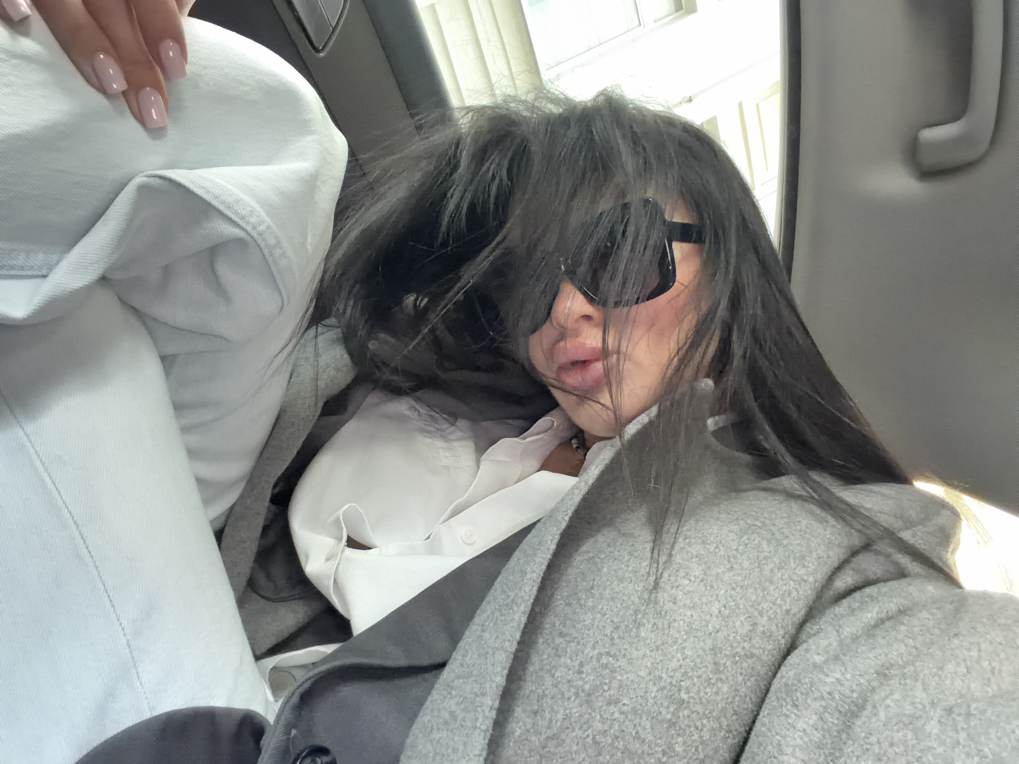 This image depicts a woman inside a car, presumably in Moscow, Russia, captured from a slightly high angle. The woman, positioned on the right side of the frame, has long, dark hair that is disheveled and largely obscures her face, cascading around prominent dark sunglasses. Her lips are visible beneath the sunglasses, pursed in a slight pout.
She is wearing a light grey, textured jacket or coat over a white collared shirt. On the far left, a hand with neatly manicured, light pink nails gently rests on a large pile of light blue fabric, possibly denim, which appears to be folded or bundled in the car's interior. Additional white and dark grey fabrics are visible in the background, suggesting more clothing or soft items piled around her.
The setting is clearly inside a vehicle, with parts of the car's interior visible, including a grey ceiling handle on the right and what appears to be a door or pillar. Through the windows, indistinct shapes of buildings or structures under natural daylight suggest an urban environment during the daytime. The woman appears to be posing casually for a photograph or selfie, her expression conveyed through her lips and the overall stylish yet messy aesthetic. There is no visible text in the image.
