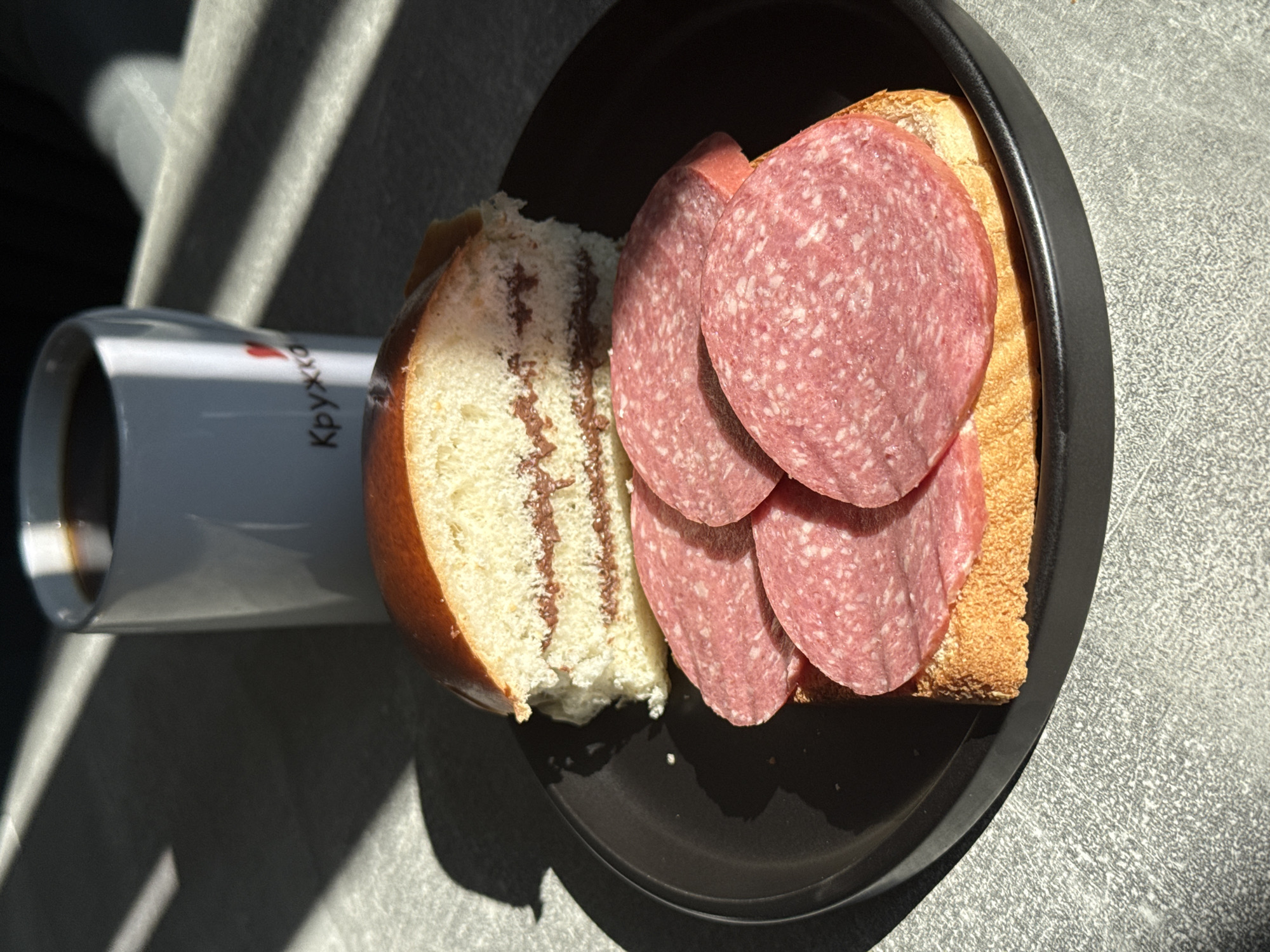 The image shows a close-up of a breakfast scene. In the foreground, on a black plate, is a sandwich on a slice of toasted bread topped with three slices of salami. Behind the sandwich, on the plate, is a halved brioche bun filled with a chocolate spread. In the background, a white mug filled with dark liquid, likely coffee, is partially visible, with the word "Кружка" (Kruzhka, meaning mug in Russian) and a heart symbol printed on it. The scene is illuminated by sunlight casting strong shadows from what appears to be blinds or a window. The location is the city of Lyubertsy, Russia.