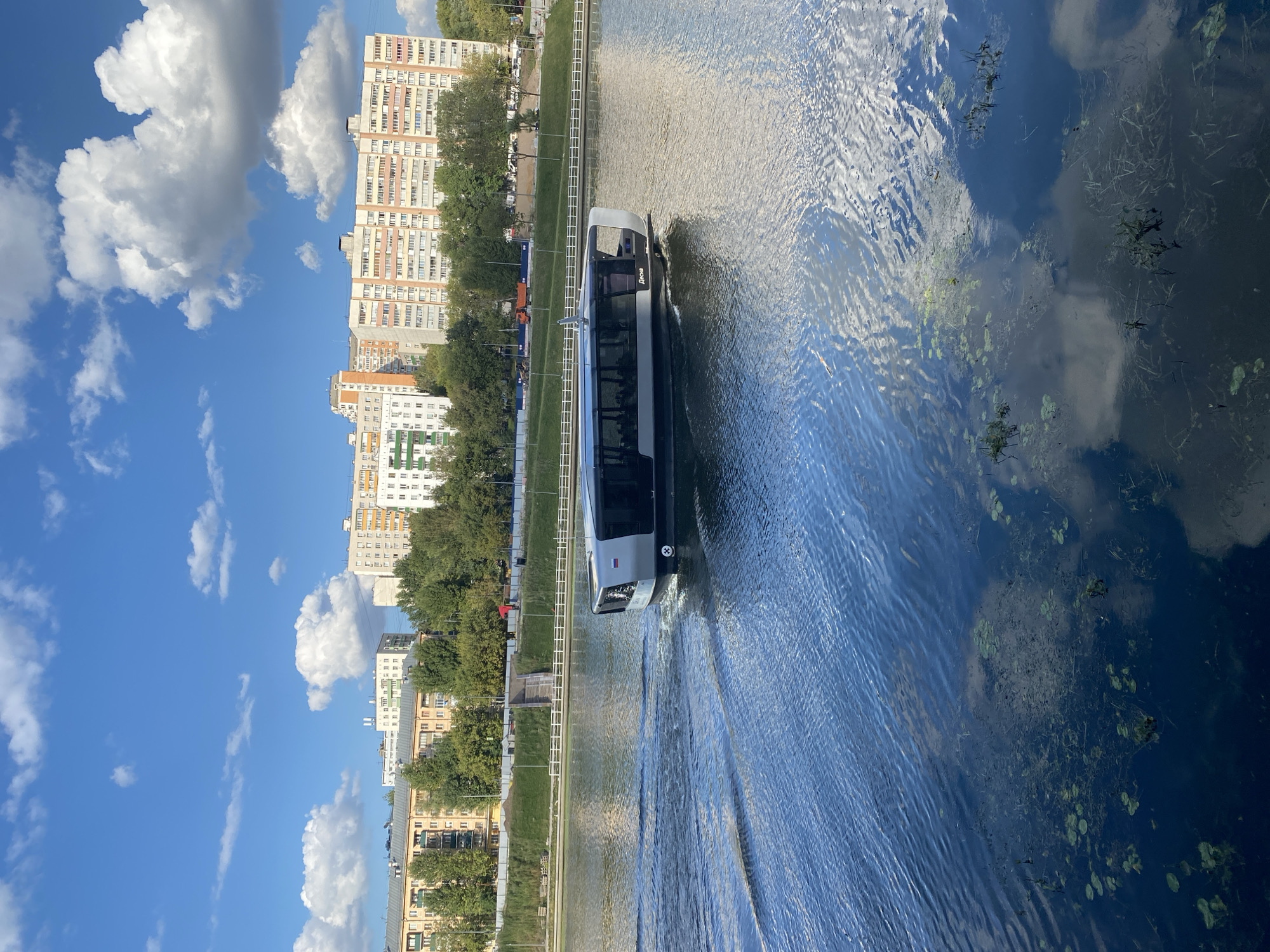 An eye-level, medium shot shows a modern, gray and black passenger boat with large windows moving across a body of water. The boat is situated on the right side of the frame, creating a wake behind it. Reflected in the water are clouds from the bright blue sky. In the background, a green embankment with trees and a staircase leads up to a series of tall apartment buildings. The scene appears to be a calm, sunny day in a city.