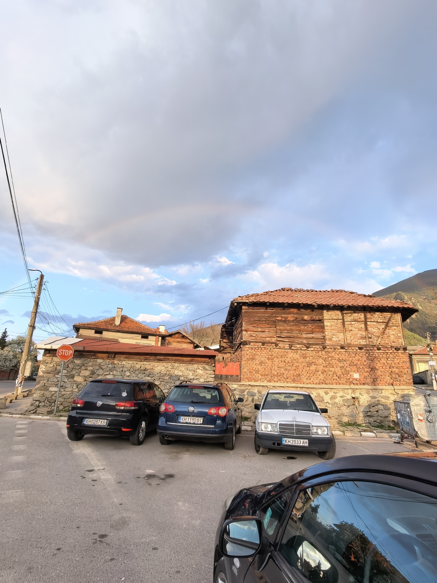 The image shows a street scene with three parked cars in front of a stone and brick building in Unknown, Unknown. A black Volkswagen Golf with license plate KH0287KB, a blue Volkswagen Passat with license plate KH1190BT, and a white Mercedes-Benz with license plate KH2033AH are parked along the street. The building behind the cars is constructed with a stone base, brick walls, and a wooden upper section with a tiled roof. A stop sign is visible to the left of the cars. In the background, there are additional buildings and trees. The sky is partly cloudy, and a faint rainbow is visible in the upper left portion of the image. A dark-colored car is partially visible in the foreground.