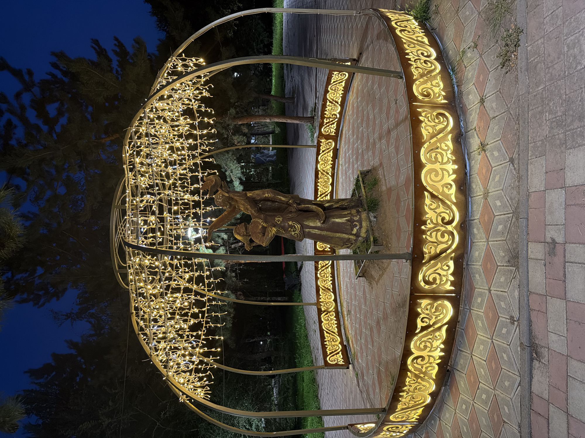 This image shows a bronze statue of two figures within a circular, illuminated structure in a park at night. The structure is supported by metal posts and features a decorative border with ornate patterns illuminated by warm, golden lights. The ceiling of the structure is adorned with numerous small, dangling lights, creating a canopy effect. The statue appears to depict a man and a woman in traditional attire, with the woman holding a bird. The ground within the structure is paved with diamond-shaped tiles in red and beige. Surrounding the structure, the park features grass, trees, and pathways, also illuminated by park lighting in the background. The sky is dark blue, indicating nighttime. This scene is located in the City Park of Pavlodar, Kazakhstan.