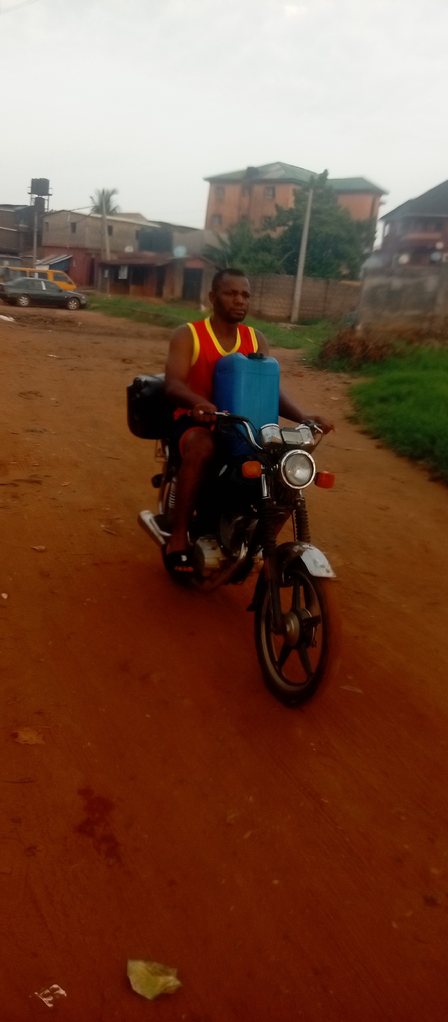 In Ogidi, Nigeria, an individual is riding a black motorcycle down a red dirt road. The rider wears a red and yellow sleeveless top and dark shorts. A blue plastic container sits in front of him, and a black bag is mounted on the rear of the bike. The environment features buildings, vegetation, and cars visible in the background under an overcast sky.