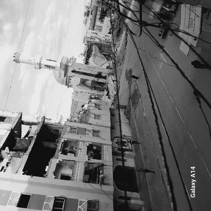 This black-and-white image, captured from an elevated perspective, displays a scene of widespread structural damage in the city of Khan Yunis, Palestinian Territory, under an overcast sky. Numerous dark, tangled cables crisscross the upper foreground. To the left, a multi-story building, likely residential, exhibits severe damage with missing exterior walls exposing several internal floors and debris on balconies. To the right, a prominent mosque with a tall, slender minaret and a dome is heavily damaged, its facade partially collapsed, and extensive rubble is strewn around its base. The asphalt street below features white lane markings and is flanked by piles of debris. Four individuals are visible: one person in light clothing walks left on the far-left sidewalk, another person in dark clothing walks right on the left sidewalk closer to the camera, a person in dark clothing walks away from the camera in the middle of the street, and a fourth person in dark clothing is partially obscured near a damaged white vehicle and a banner with Arabic text on the right side of the street. Additional damaged structures extend into the background. A "Galaxy A14" watermark is present in the bottom left corner.