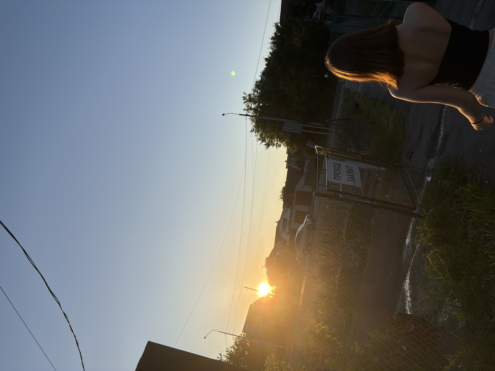 The image captures an outdoor scene at sunset, likely in Новинки, Russia, based on provided context. The sun, a bright orange orb, is low in the sky on the left, casting a strong glare and warm light across the landscape. The sky above is clear, transitioning from bright yellow near the horizon to a pale blue overhead. Multiple dark power lines traverse the upper portion of the sky, with a notable green circular lens flare visible in the upper right.
In the foreground, a chain-link fence runs horizontally, separating an overgrown grassy area from a paved path or road beyond. Attached to the fence on the right is a white sign with black Cyrillic text reading "ПРОХОД ЗАКРЫТ" (PASSAGE CLOSED), indicating a restricted area.
A sole individual is visible from behind in the lower right corner, walking away from the camera towards the closed gate. This person has short, reddish-brown hair, illuminated by the direct sunlight, and wears a black strapless top with light-colored bottoms. A dark band is present on their left wrist.
Beyond the fence and path, several residential buildings with varied rooflines are silhouetted against the setting sun. A light-colored sedan is visible on the road in front of these buildings. A streetlight pole stands to the left of the path, and another taller streetlight is visible further right, partially obscured by a large, dark-leafed tree. On the far left, a partially visible sign with "OZON" and "WU" is discernible on a building. The immediate foreground features dense, sun-drenched vegetation.