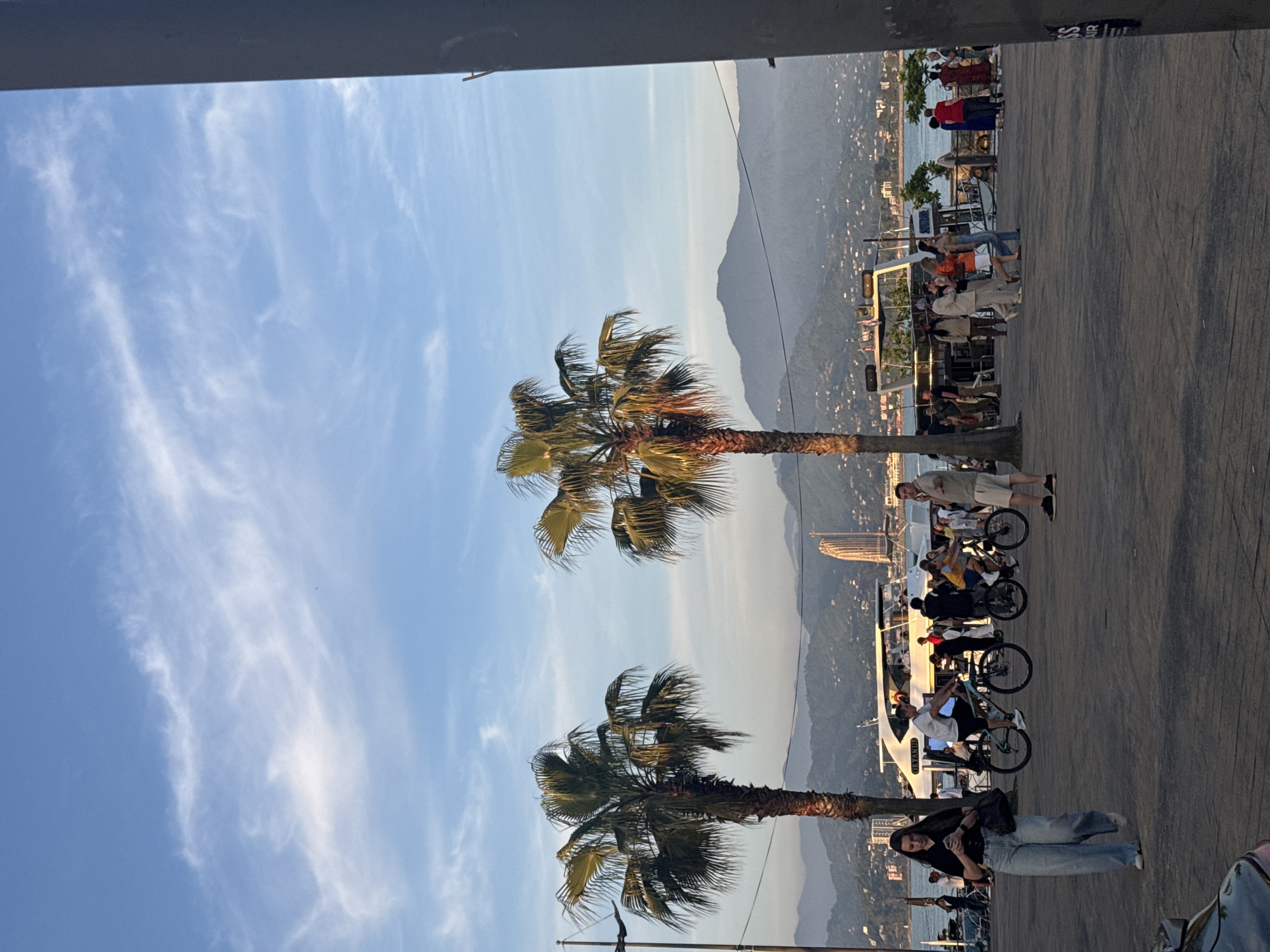 The image depicts a vibrant waterfront scene in Batumi, Georgia, during the late afternoon or golden hour, characterized by warm, soft sunlight. The sky is a clear blue with scattered, wispy white clouds. In the foreground, a paved promenade features several individuals. A young woman in a dark top, light-wash wide-leg jeans, and white sneakers walks towards the left, looking down at her smartphone while carrying a dark shoulder bag. To her right, a man in a white t-shirt and dark shorts rides a bicycle to the right, followed by another cyclist. Further right, a man in a light t-shirt and shorts stands, looking towards the right with his left arm slightly raised.
Two prominent palm trees with dense green fronds, illuminated by the warm sunlight, frame the midground. Behind the people and palm trees, a body of water, likely the Black Sea, is visible. Several boats are docked or cruising; a large white yacht with numerous individuals on board is positioned centrally, and a smaller vessel with a canopy is to its right.
The background features a city sprawling across a coastal plain and extending up into distinct, hazy mountains. A tall, distinctive spiraled or twisted skyscraper, recognizable as a landmark in Batumi, stands prominently on the distant shore, slightly to the left of the center. Other buildings and urban infrastructure are visible along the coastline and hillsides. The far-right edge of the image is partially obstructed by a dark, vertical structural element. A utility pole with wires is present on the far-left.