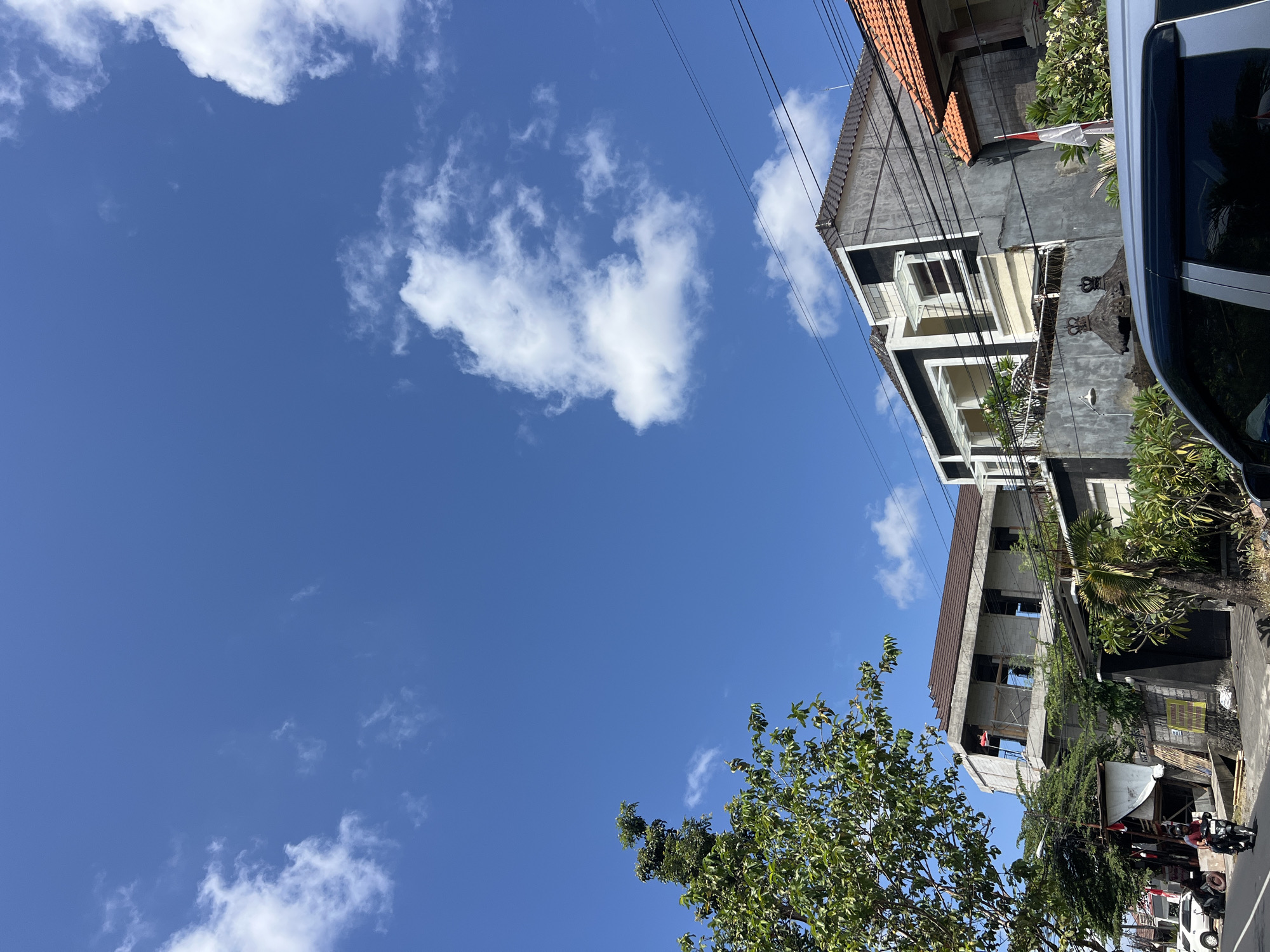 The image depicts a street scene in Denpasar, Indonesia, under a bright blue sky with scattered white clouds. In the foreground, a silver car's windshield and roof are visible, partially obstructing the view of the street. A person is riding a motorcycle on the road, heading towards the left side of the frame.
To the right of the road, several buildings are present. A partially constructed concrete building with exposed rebar and unfinished walls occupies the center-left portion of the frame, topped with a brown tiled roof. Adjacent to it, to the right, is a finished modern building with a dark gray facade, white trim around the windows, and a lighter colored section below. This building has a balcony on the second floor. Further to the right, a building with orange tiled roofing is visible, with traditional Balinese architectural elements such as wooden pillars and thatched roofs visible at the base. A red and white Indonesian flag is attached to a pole in front of this building.
Vegetation, including trees with green leaves, is present throughout the scene, some growing along the street and others around the buildings. Power lines crisscross the sky above the buildings. The overall impression is a view of a street in a tropical urban environment, likely during daytime.