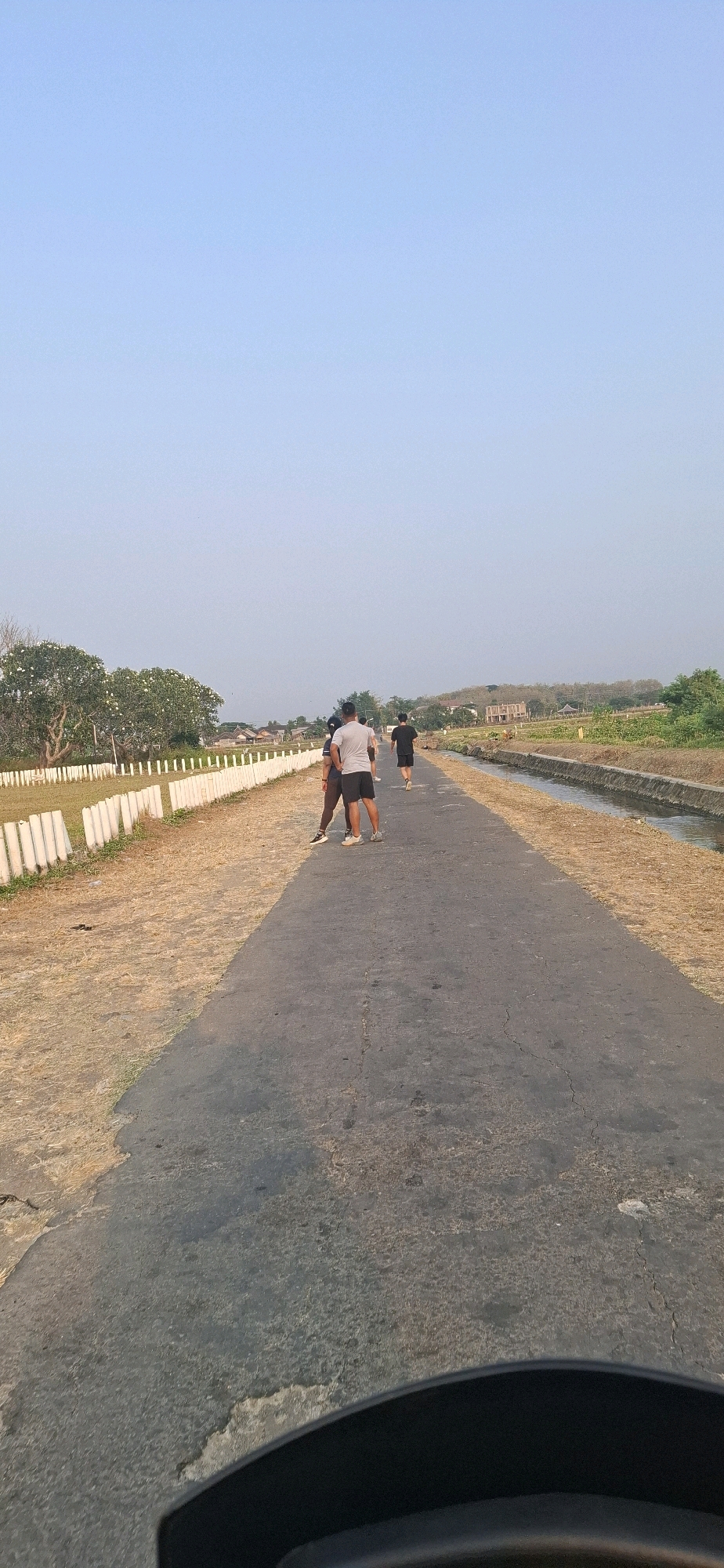 In Desa Ngesrep, Indonesia, on a paved road with dry grass and a narrow irrigation ditch to the right, three individuals are visible. One person in a white t-shirt and black shorts is standing in the middle of the road, facing away from the camera. To their left and slightly behind, another person in a dark t-shirt and shorts is also standing. Further down the road and to the right, a third person in a black t-shirt and shorts is running away from the camera. A white fence borders a grassy area on the left side of the road. In the background, there are scattered houses and trees under a clear, pale blue sky. The foreground shows a portion of a black object, possibly part of a vehicle.