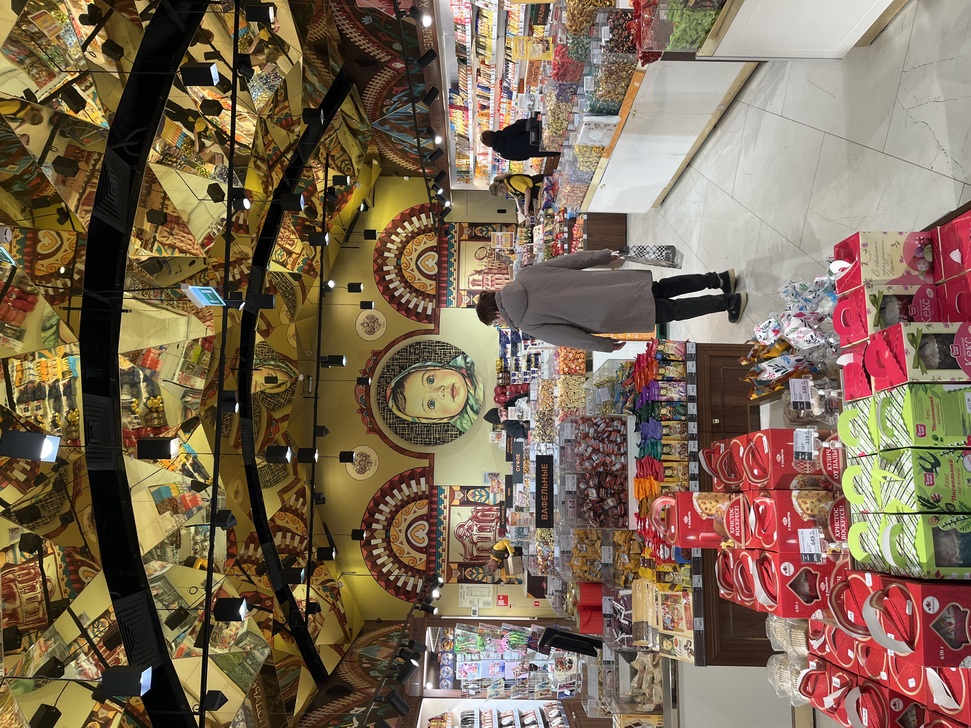 The image shows the interior of Eliseevsky Shop, a well-known grocery store in Moscow, Russia. The store's ornate ceiling with triangular mirrored panels and integrated lighting fixtures is visible above. Below, a customer in a gray coat stands in an aisle browsing products. The shelves and displays are filled with various candies, cakes, and packaged goods, including those for Easter with labels in Russian. Two other people are visible further down the aisle, one wearing a black jacket and the other a yellow shirt. The floor is tiled with a light-colored material.