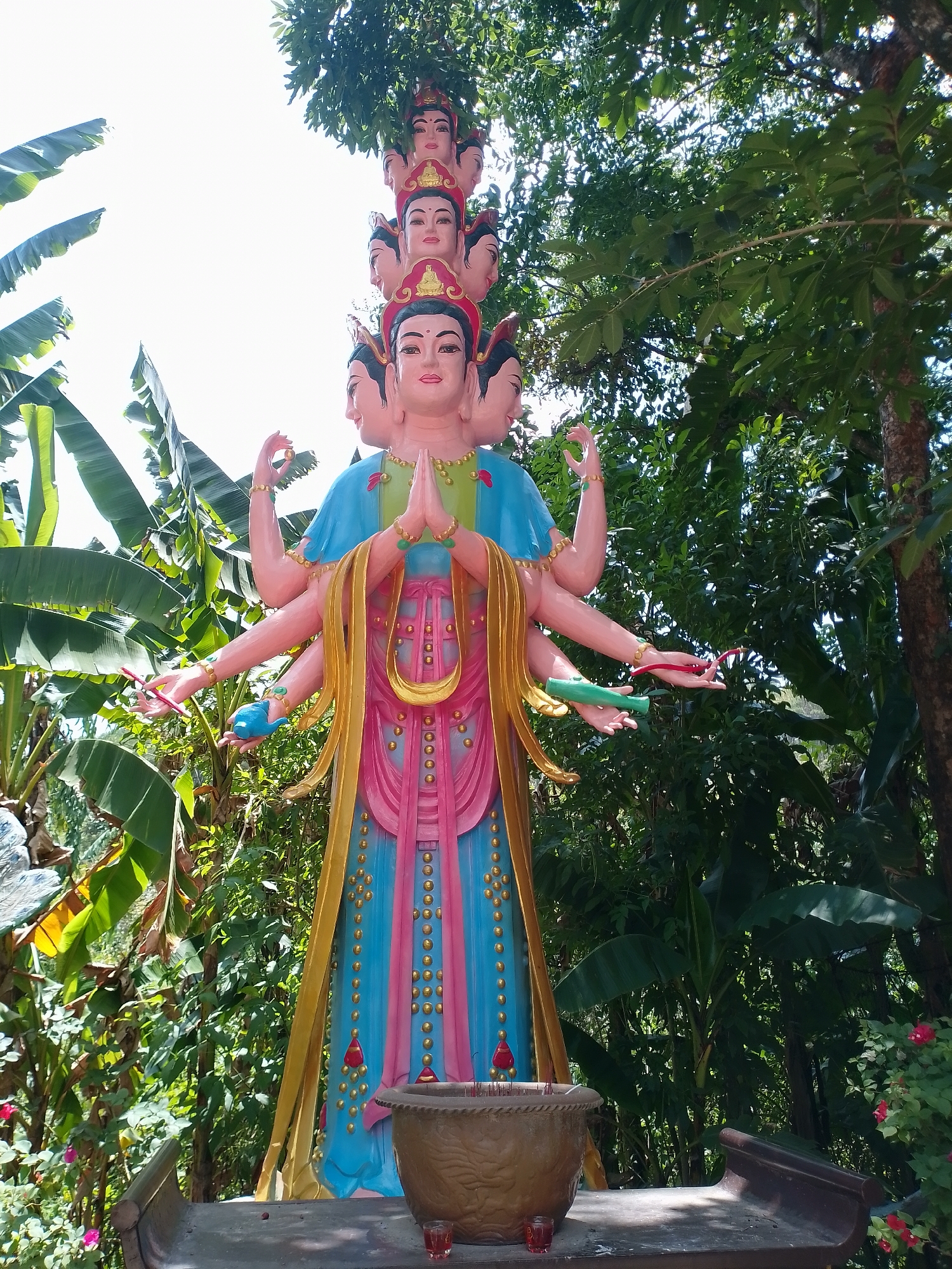 The image depicts a colorful statue of a multi-headed and multi-armed deity, likely a representation of Guanyin, located outdoors in a garden setting. The statue is adorned with various shades of pink, blue, and gold. Several heads are stacked atop the primary head, each wearing a red headdress. The deity's numerous arms extend outwards, with hands holding symbolic objects such as a writing instrument, a blue bird, a red rope, and a green tube. The statue stands on a low platform in front of a large pot, likely an incense burner. Lush greenery surrounds the statue, with banana leaves and flowering plants visible. This is located in Nam Ban, Vietnam.