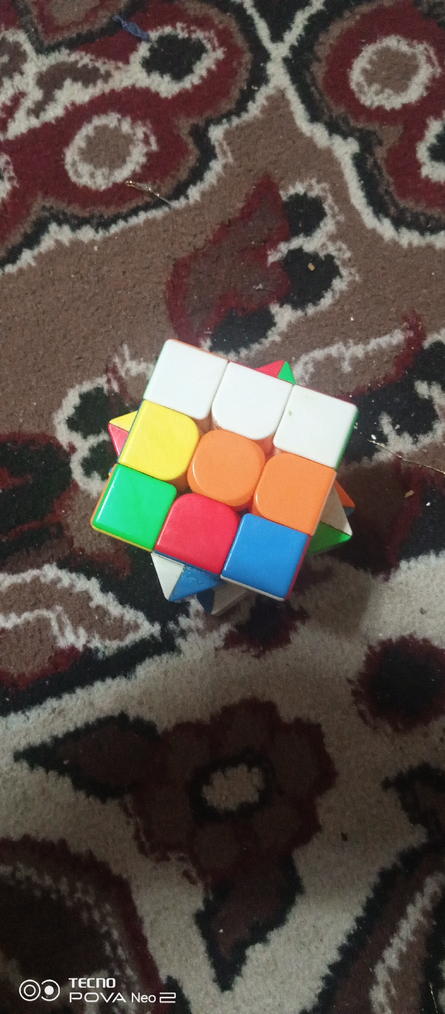 A colorful Rubik's cube is positioned on a carpet with a patterned design. The cube shows a mix of solved and unsolved sides, with colors including white, yellow, orange, green, red, and blue. The carpet has a swirling pattern in shades of brown, red, black, and white, possibly indicative of a traditional Uzbek design given the location context of Tahmach, Uzbekistan. The lighting suggests it is daytime, and no specific weather conditions are discernible. The scene is static, focusing solely on the object and its surroundings. The image also contains visible text at the bottom: "TECNO POVA Neo 2".