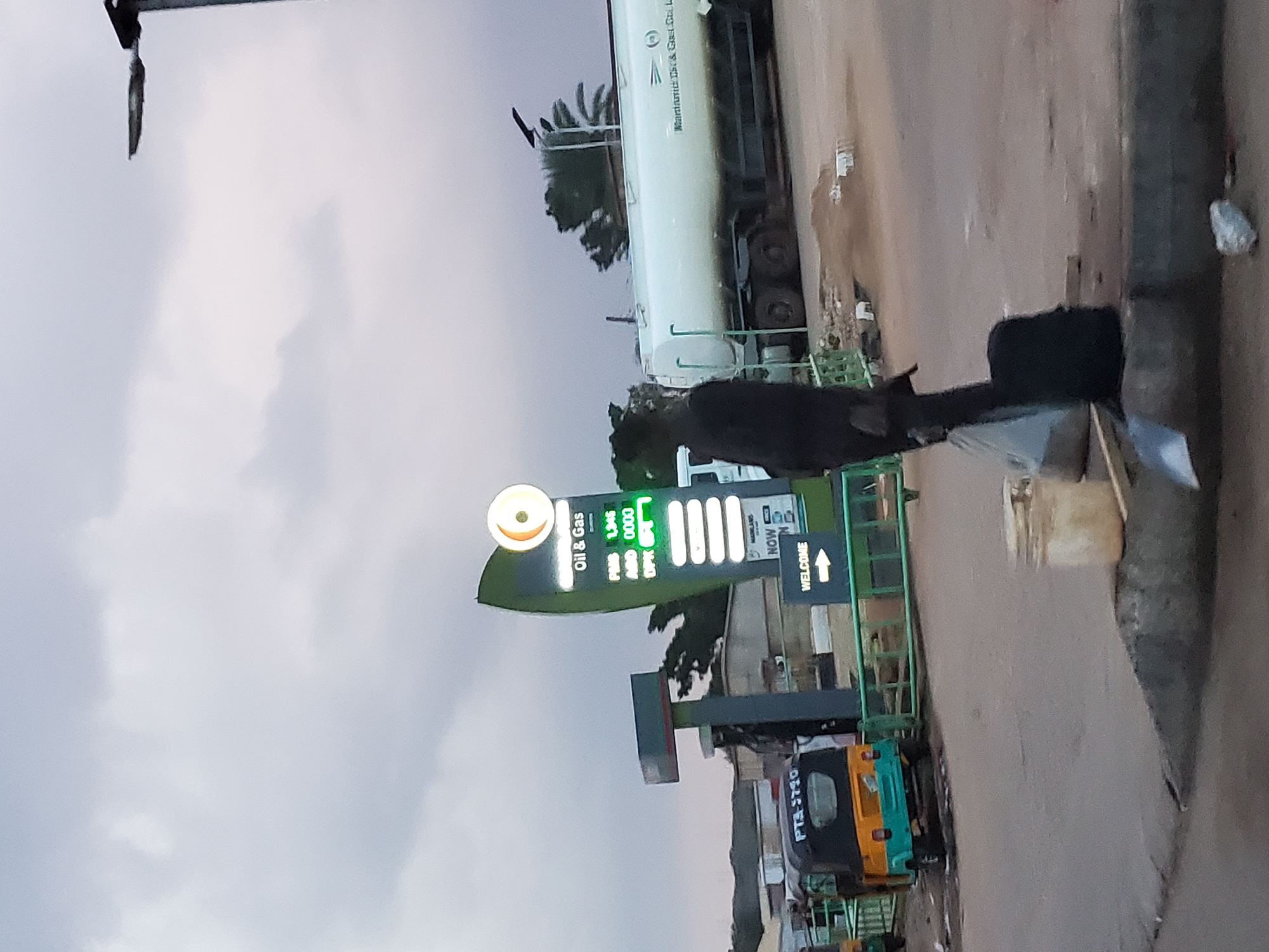 In Gura Top, Nigeria, a man in dark clothing stands near a gas station sign. The sign displays "Oil & Gas" and fuel prices, with "PMS 1,345" and "DPX 0000" visible. A white tanker truck with the company name "Mainland Oil & Gas Co. Ltd." is parked in the background. A yellow and green auto-rickshaw with the license plate "PTS-3710" is parked on the left. A streetlight is visible in the upper right corner. The scene appears to be an outdoor setting with a cloudy sky.