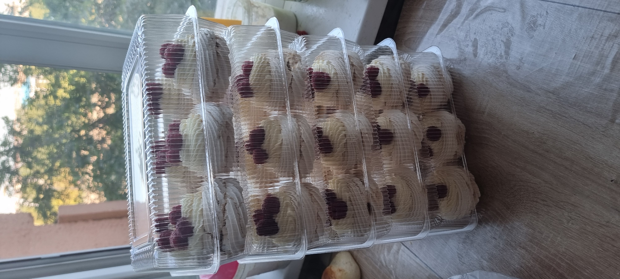 The image displays a stack of clear plastic containers filled with individual meringue desserts topped with white frosting and raspberries. The containers are placed on a wooden surface, likely a table or countertop, near a window with blurred greenery visible outside. A small, golden-brown pastry is partially visible on the left side of the image, and some containers and jars are visible in the background. The scene appears to be indoors, with natural light coming from the window. There are no people visible in the image. The desserts are arranged in multiple layers within the containers, with each dessert positioned in its own compartment. The overall impression is one of a prepared batch of desserts, possibly for sale or a special occasion. The location is identified as Aktobe, Kazakhstan.