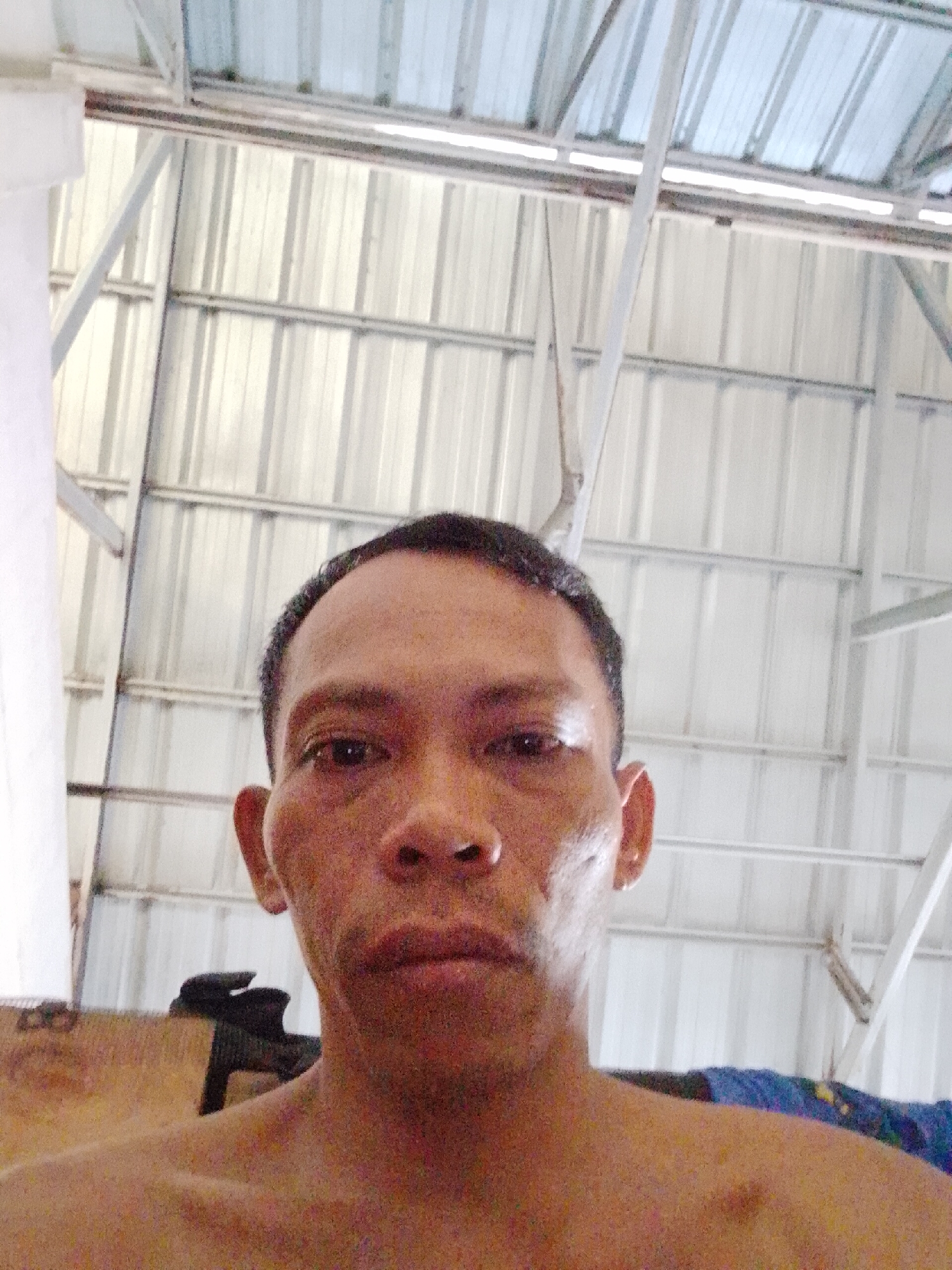 A close-up, eye-level selfie of a man with dark, short hair. The man is facing the camera with a neutral expression. The background consists of a metal corrugated roof supported by a metal frame. The photograph appears to have been taken indoors. The location is specified as Bukit Ketok, Indonesia, but no evidence within the image confirms this.