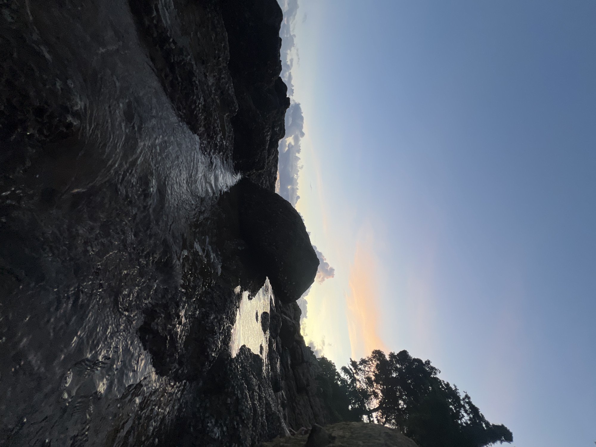 The image depicts a natural coastal scene at dusk. The primary subjects are the rough, dark rocks that line a narrow body of water, likely a tidal pool or a small stream. A large, dark rock juts out into the water, casting a shadow. The water itself is dark and reflects the subdued light of the setting sun, creating shimmering patterns on its surface and on the wet rocks.
In the background, silhouetted trees are visible against a sky that transitions from a pale, warm orange near the horizon to a clear, deep blue overhead. Scattered clouds with soft edges are present in the mid-sky, illuminated by the fading sunlight. The overall mood is serene and tranquil, characteristic of the end of the day.
The time of day is clearly evening, indicated by the soft lighting and the colors of the sky. There are no people or discernible human-made objects, emphasizing the natural beauty of the location. The rocky terrain and the presence of trees suggest a coastal or rugged landscape. The hints provided indicate the location is Choeng Thale, Thailand, which is consistent with a tropical coastal environment. There is no visible text in the image.