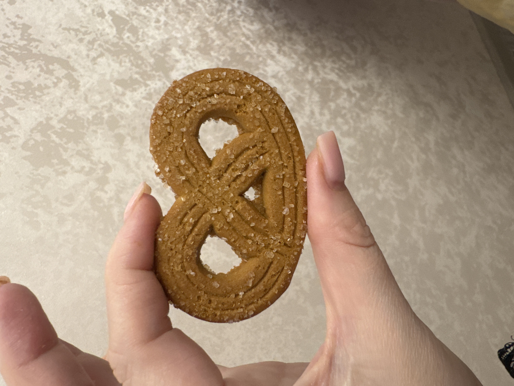 A close-up shot features a light brown, pretzel-shaped cookie, generously coated with coarse sugar crystals, held by a person's hands. The cookie is the central focus, showcasing its twisted, figure-eight design and sparkling sugary texture. The hands, with short, natural-looking nails, grasp the cookie from either side, presenting it to the viewer.
The background is a light, off-white surface with a subtle, irregular, and textured pattern, resembling a marbled countertop or tablecloth. Soft, ambient lighting illuminates the scene, casting gentle shadows on the background. No specific time of day or emotional state is discernible. There is no visible text. This indoor scene is located in Beryozovy, Russia.
