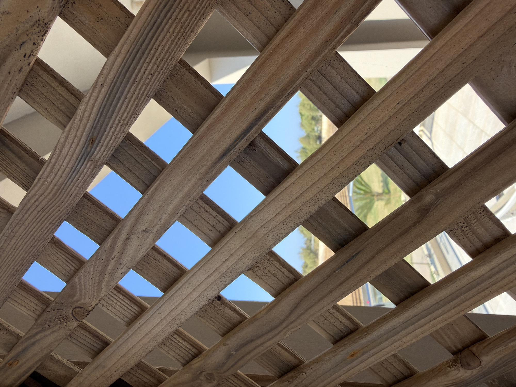 The image shows a close-up of a wooden lattice with a diamond pattern, viewed from below. Through the openings in the lattice, a bright blue sky is visible, along with hints of distant greenery and architectural structures. The wood appears weathered and has a natural grain. The perspective suggests the viewer is looking upwards through the lattice, possibly from a patio or balcony. The location is identified as Vigklapia, Greece.