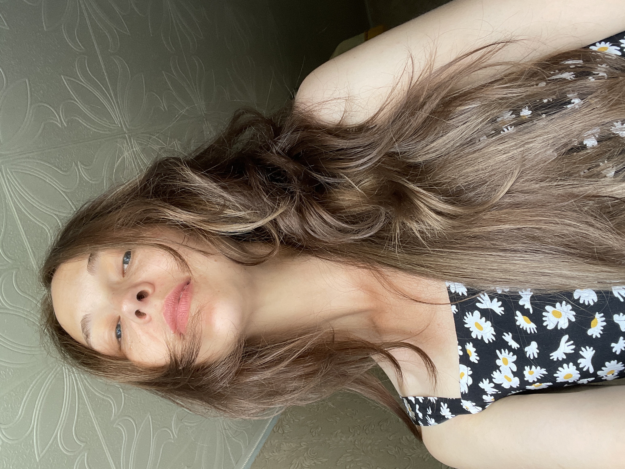 A low-angle, medium shot shows a woman looking up towards the camera. She has long, light brown hair that is slightly messy and falls around her face and shoulders. Her eyes are blue and her lips are slightly parted, showing a hint of pink lipstick. She is wearing a black tank top with a white daisy pattern. The background consists of a textured ceiling and a wall with a floral pattern wallpaper. The lighting appears to be natural and coming from above.