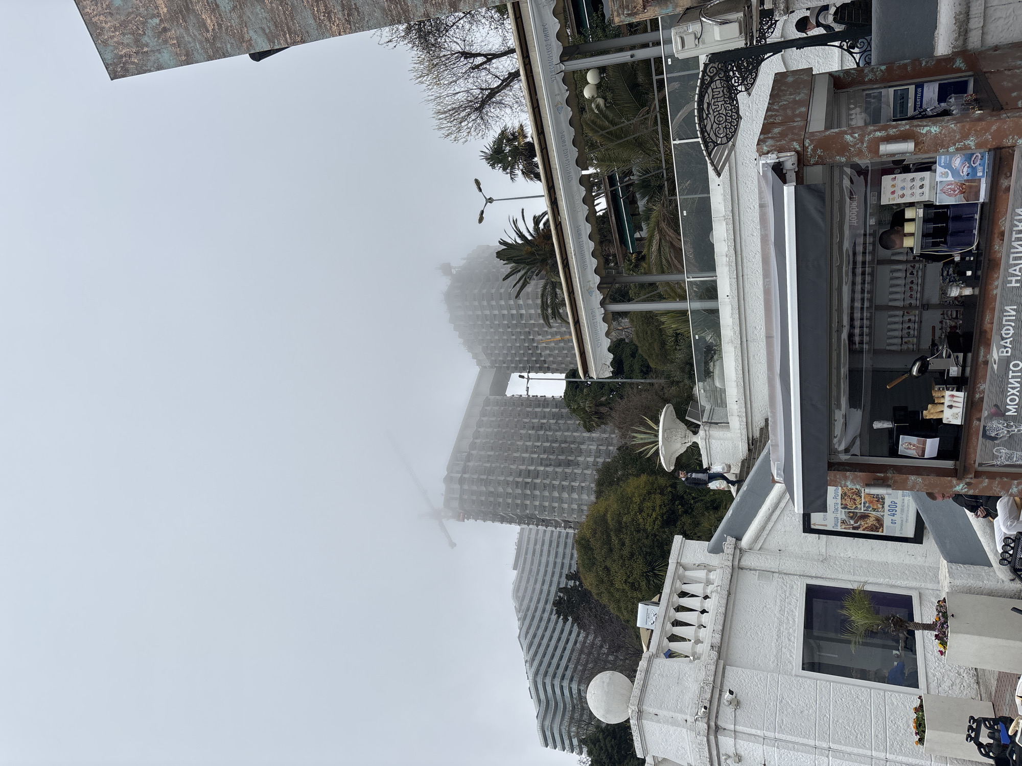 This outdoor shot captures a foggy day in Sochi, Russia. The scene showcases a multi-storied building under construction, partially obscured by fog, behind a smaller white building with a kiosk.
The kiosk, with signage in Russian, displays an image of pizza. A few people are visible near the kiosk, and a potted plant can be seen in a nearby window. The architectural style is reminiscent of older resort towns, and palm trees suggest a subtropical climate.