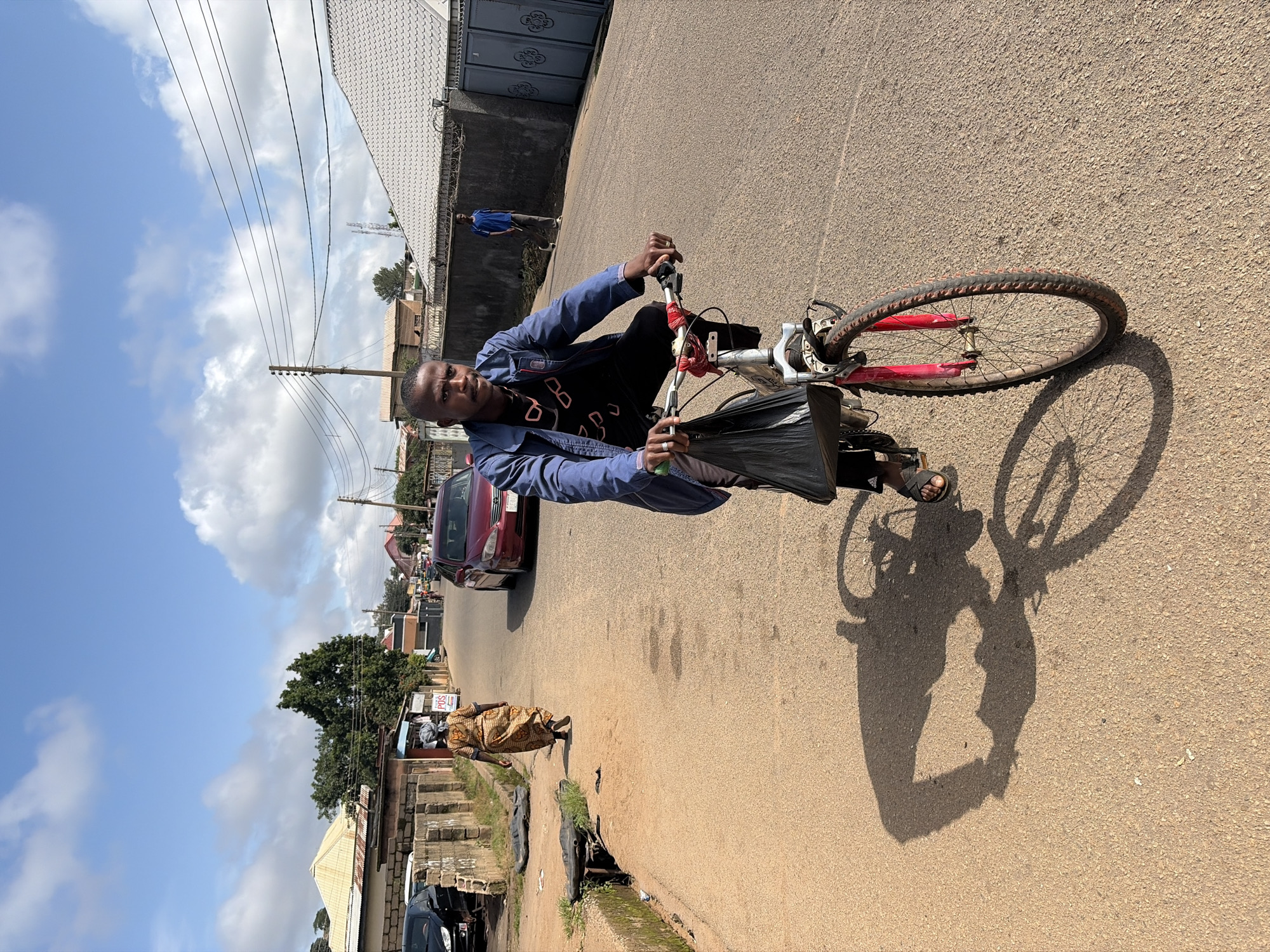 A medium shot captures a man riding a bicycle down a sunlit, paved street in Jos, Nigeria. The man, with dark skin and a close-cropped haircut, wears a blue jacket over a black t-shirt and black pants, and sandals on his feet. He is holding black handlebars and carrying a black plastic bag in front of him on the bicycle. The bicycle has a silver frame, red rims, and a red bandana tied to the handlebars. A large shadow of the man and his bicycle is cast on the asphalt road. In the background, a woman in a patterned golden dress walks away from the camera on the left side of the street, her head covered. Further down the street, a red car is driving away, and a young boy in a blue t-shirt and gray pants walks towards the camera on the right side of the street. The street is lined with buildings, fences, and vegetation. The sky above is blue with scattered white clouds. A power line runs across the upper portion of the frame.