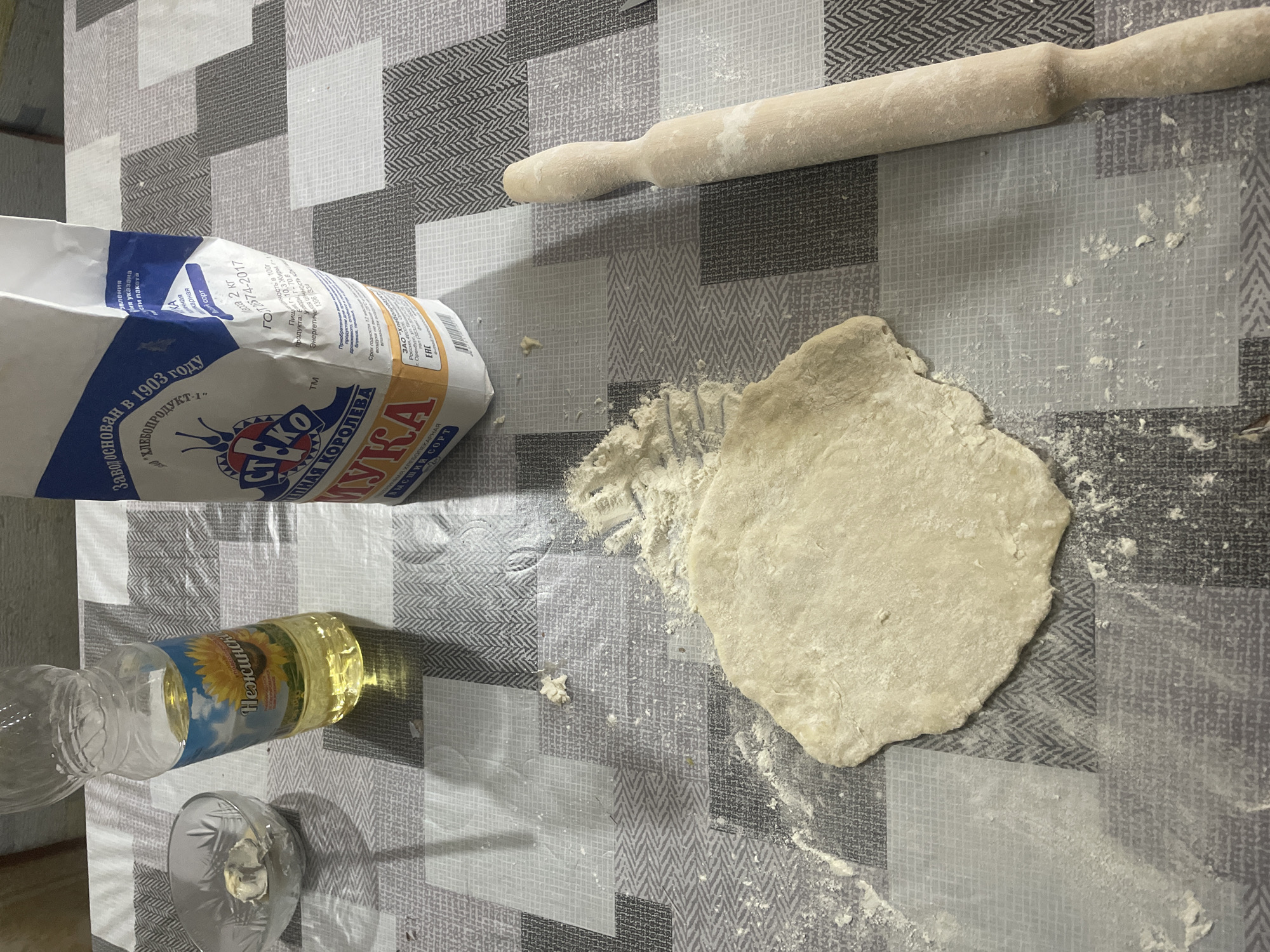 The image shows a table covered with a patterned tablecloth, where dough is being prepared. Scattered across the table are several objects related to baking. In the upper right corner, a wooden rolling pin lies horizontally. To the left of the rolling pin, a flattened disc of dough is visible, dusted with flour. Next to the dough, a bag of flour is partially visible, with text in Russian, including "ХЛЕБОПРОДУКТ-1" and "Мука". In the lower left corner, a partially empty plastic bottle of sunflower oil with a label featuring a sunflower and Russian text "Нежинская" is placed horizontally, next to a small glass with some liquid. The overall scene suggests the preparation of some baked goods, likely bread or pastries, in a domestic setting. The lighting is moderate, suggesting an indoor environment, possibly during the daytime. The background tablecloth has a geometric pattern of squares and zigzags in shades of grey and white. There are no people visible in the image, and no clear indication of the time of day or specific weather conditions.