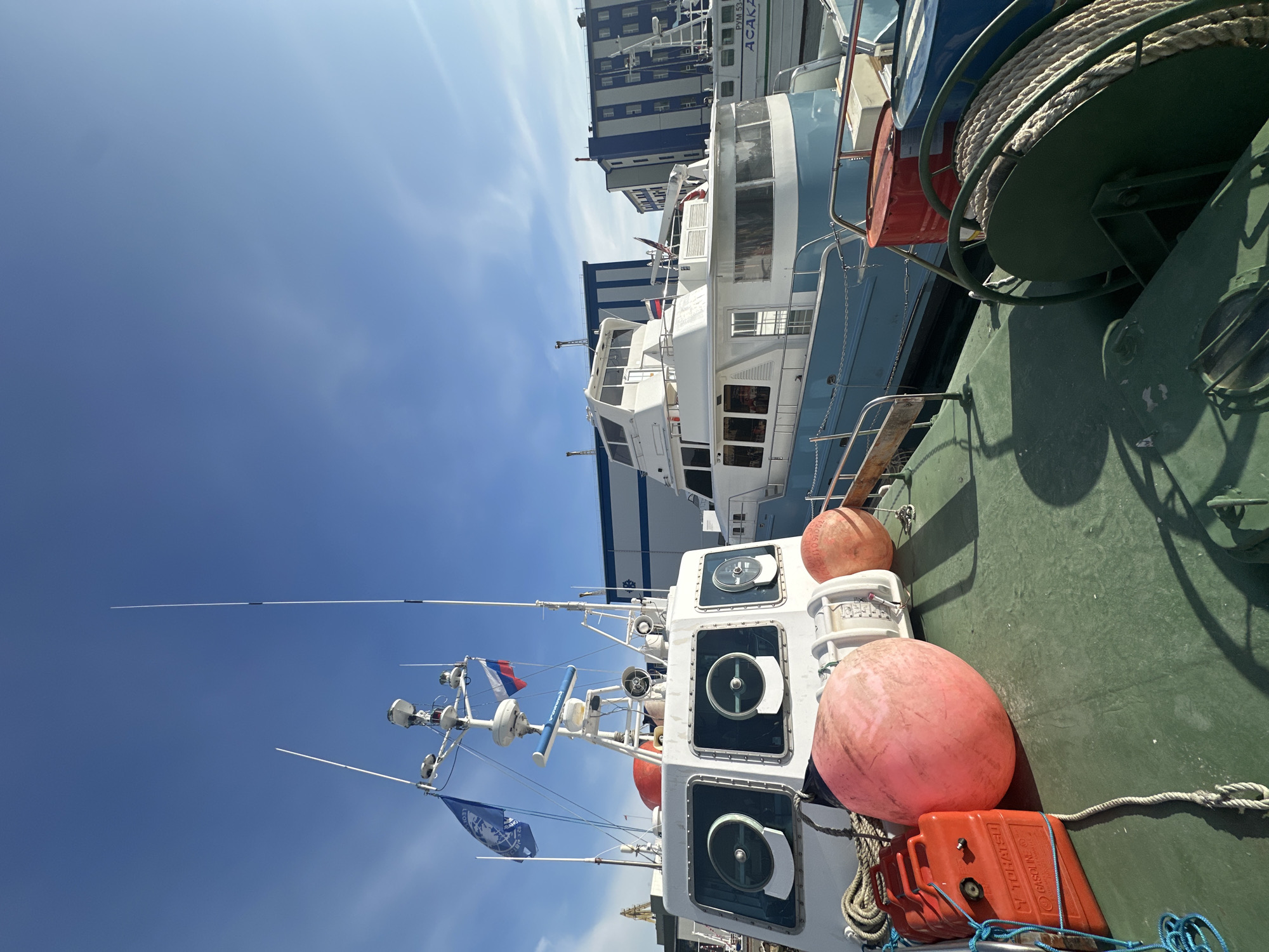 The image captures a view of docked boats in the port of Petropavlovsk-Kamchatsky, Russia, under a clear blue sky. In the foreground, a green-decked vessel is visible, adorned with large orange buoys and an orange fuel canister labeled "TOHATSU GASOLINE." Behind this vessel, a white boat with large circular windows is prominent, featuring various antennas and flags, including the Russian flag. Further in the background, a larger, light-blue and white yacht is moored alongside. Industrial buildings, painted with vertical blue and white stripes, form the backdrop. The scene is illuminated by sunlight, casting shadows across the deck.