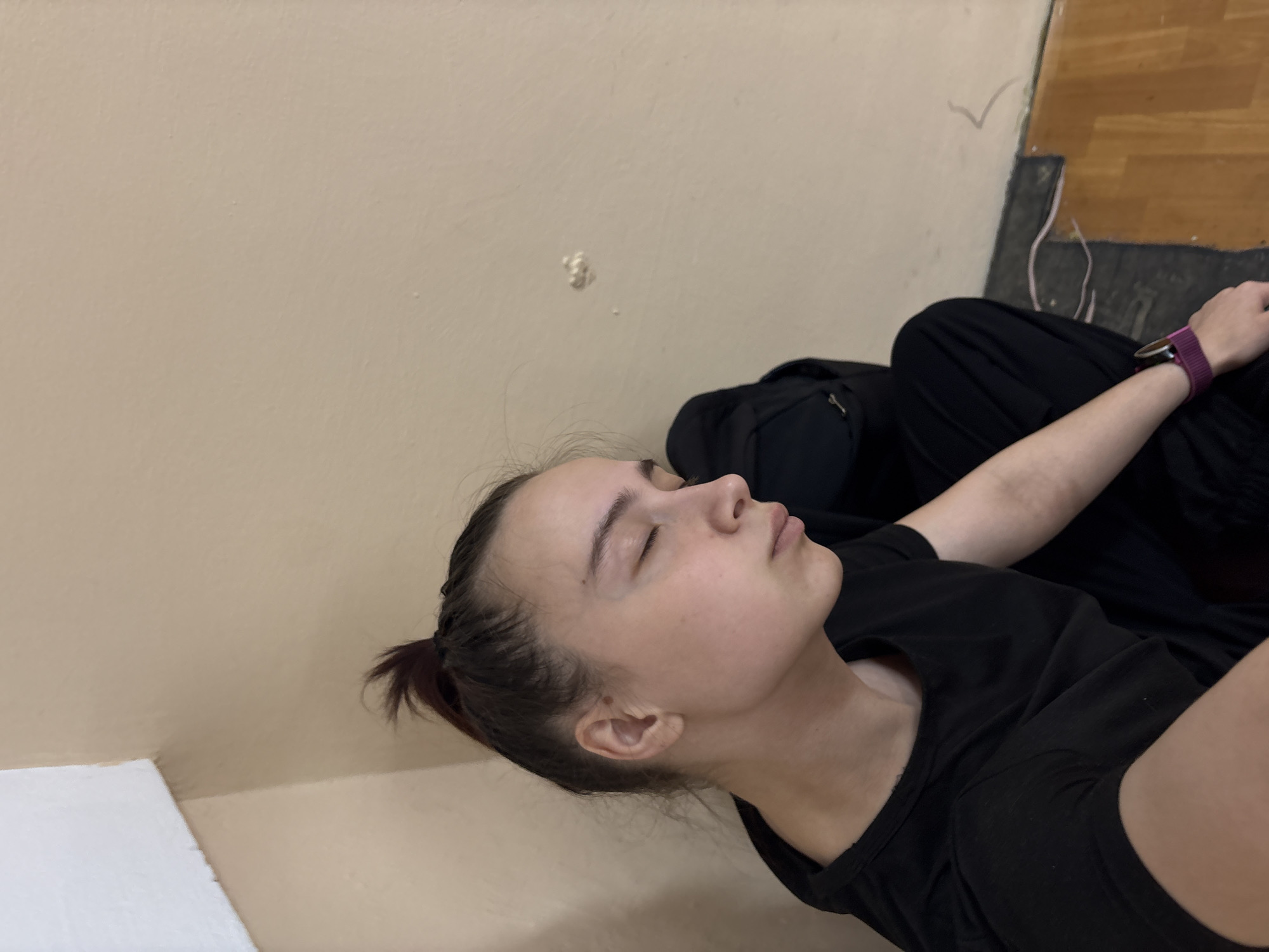 A young woman with fair skin is resting with her eyes closed. Her dark hair is pulled back in a small ponytail. She is wearing a black t-shirt and has a smartwatch with a purple band on her left wrist. The background consists of a beige wall with a small blemish or patch. A dark-colored fabric is bunched behind her, and the edge of a wooden floor is visible in the upper right corner. Based on the location, this image was taken in Chaikovsky, Russia.