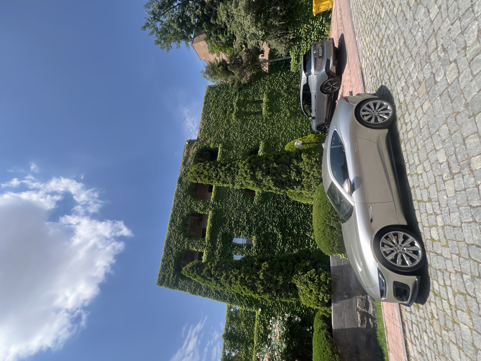 In Yerevan, Armenia, a modern building with a facade entirely covered in lush green ivy stands under a clear blue sky dotted with fluffy white clouds. Two cars are parked on a cobblestone driveway in front of the building. In the foreground, a silver Jaguar F-Type sports car is prominently displayed, its sleek design and alloy wheels visible. Behind it, a silver BMW iX SUV is parked further down the driveway. The surrounding environment features well-maintained green hedges and trees, creating a picturesque and serene setting.