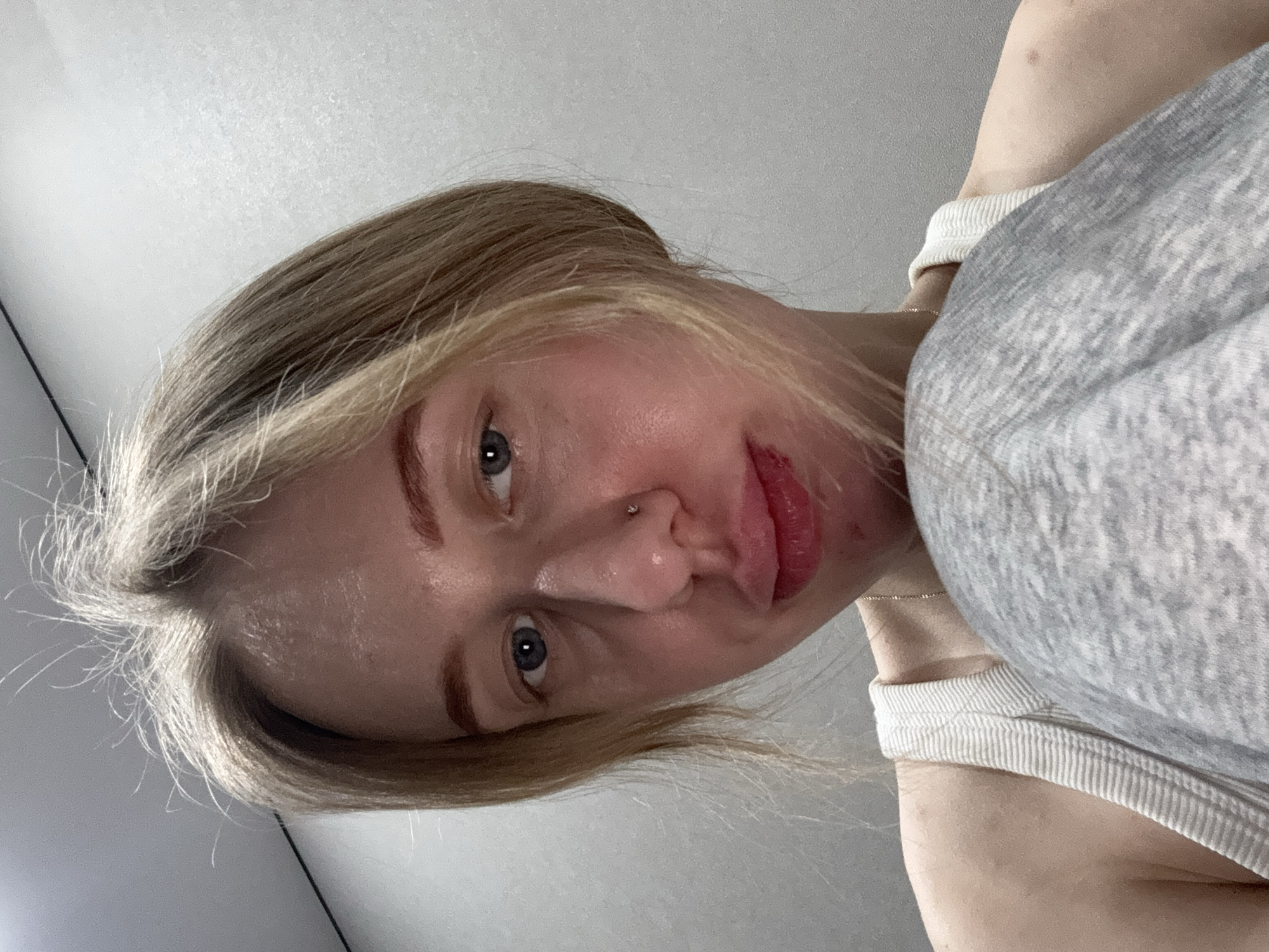 The image depicts a close-up indoor portrait of an individual, stated to be located in Rostov-on-Don, Russia. The subject is a person with fair skin, light-colored (blonde/light brown) hair, and blue eyes, looking directly at the camera. The hair appears slightly disheveled with visible flyaway strands at the crown. Subtle horizontal lines are present on the forehead. The facial skin appears somewhat reflective, particularly on the nose and cheeks. A small metallic stud piercing is visible on the left nostril. The lips are full with a reddish hue; the lower lip shows signs of redness or slight irritation. The subject is wearing a light-colored (off-white) ribbed tank top, and a thin gold chain necklace is visible around the neck. The left shoulder and upper arm are partially visible. In the foreground, a grey, mottled fabric, possibly covering a raised knee or limb, partially obscures the lower face and chest of the individual. The background consists of a plain, light grey wall, with a subtle horizontal line visible at the top left, suggesting a corner or junction with a ceiling.