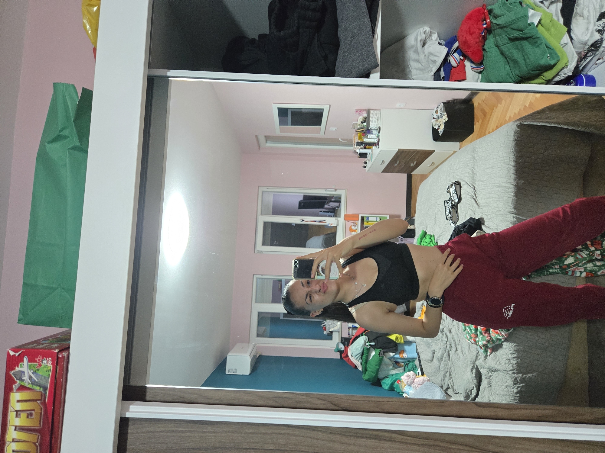 A mirror selfie shows a woman in a black sports bra and red sweatpants in a bedroom. The room has pink and blue walls, a bed with a gray comforter, and a white dresser. The closet behind her contains clothes and other items. The location is Pleven, Bulgaria.