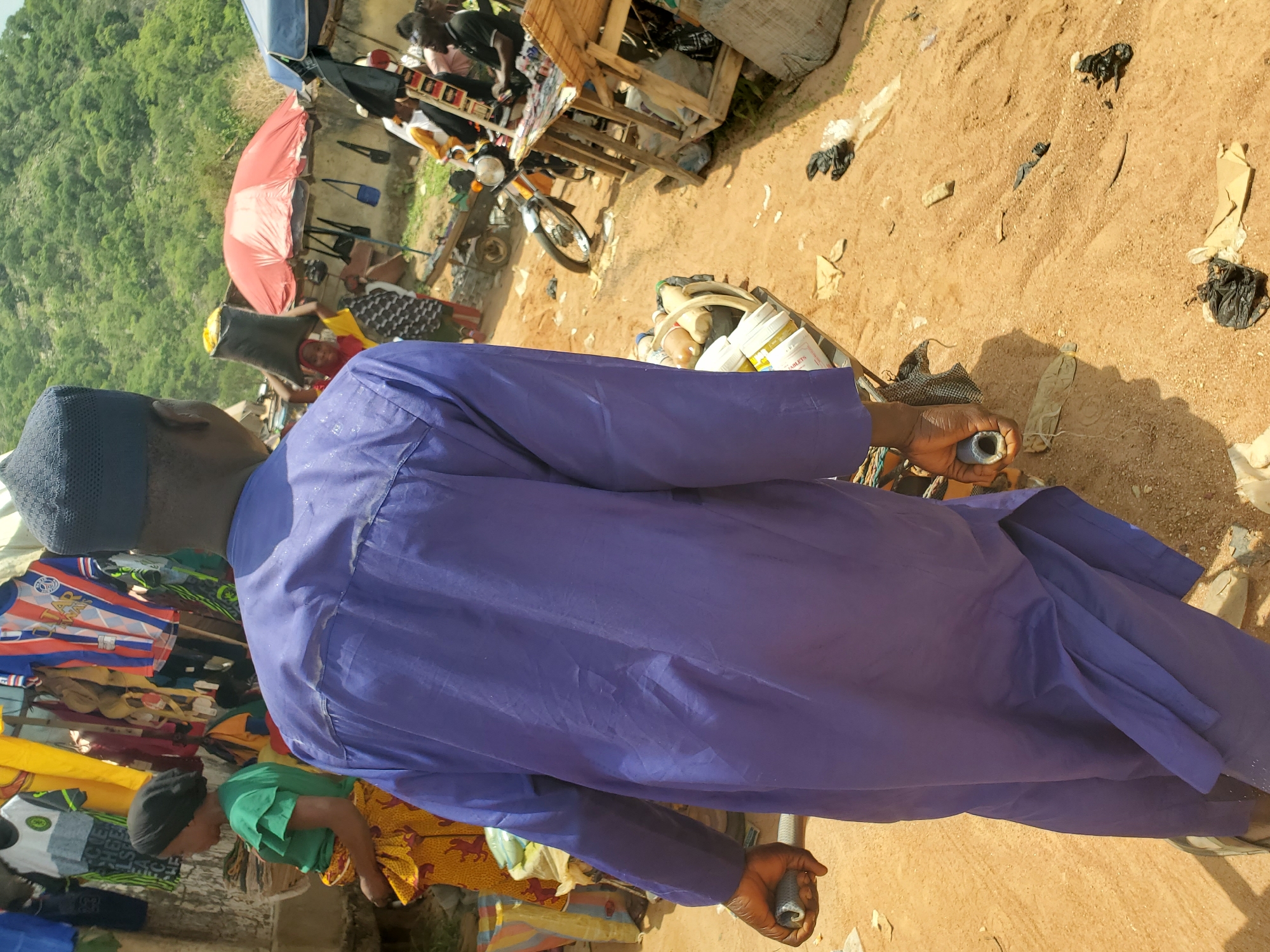 The image shows an outdoor scene in what appears to be a market setting in Mararaba, Nigeria. In the foreground, a man wearing a blue traditional garment and a dark blue cap is holding a small, metallic object in each hand. His back is facing the camera. To the left, there are clothing items displayed on a makeshift stand, suggesting a vendor's stall. Beyond the man, there are more vendors and people. A red umbrella stands out amidst the market activity. In the background, there is a hill covered with green vegetation. The ground is mostly dirt and sand, with some scattered debris. A motorcycle is visible among the vendors and merchandise.
