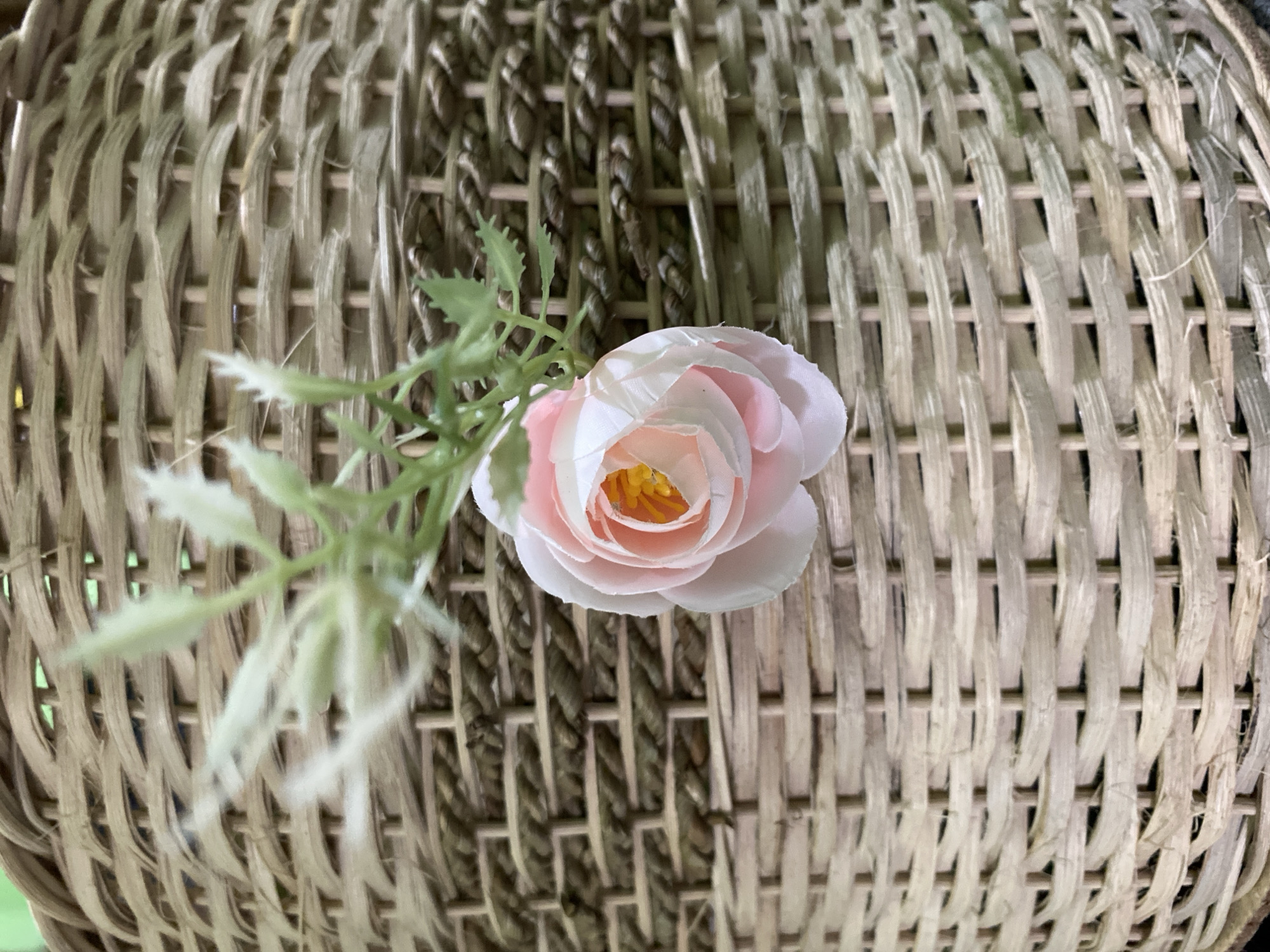 A close-up shot focuses on a light pink artificial rose with a yellow center, attached to a thin green stem with small, fluffy white and green leaves. The rose and stem are positioned on the right side of the frame, slightly off-center. The background is a tightly woven basket made of natural fibers, predominantly in beige and light brown tones, with a slightly distressed texture. The weave pattern is a consistent over-under interlacing. The lighting casts subtle shadows, highlighting the texture of the basket. There is no indication of the city of Kuala Pembuang, Indonesia, or any people.