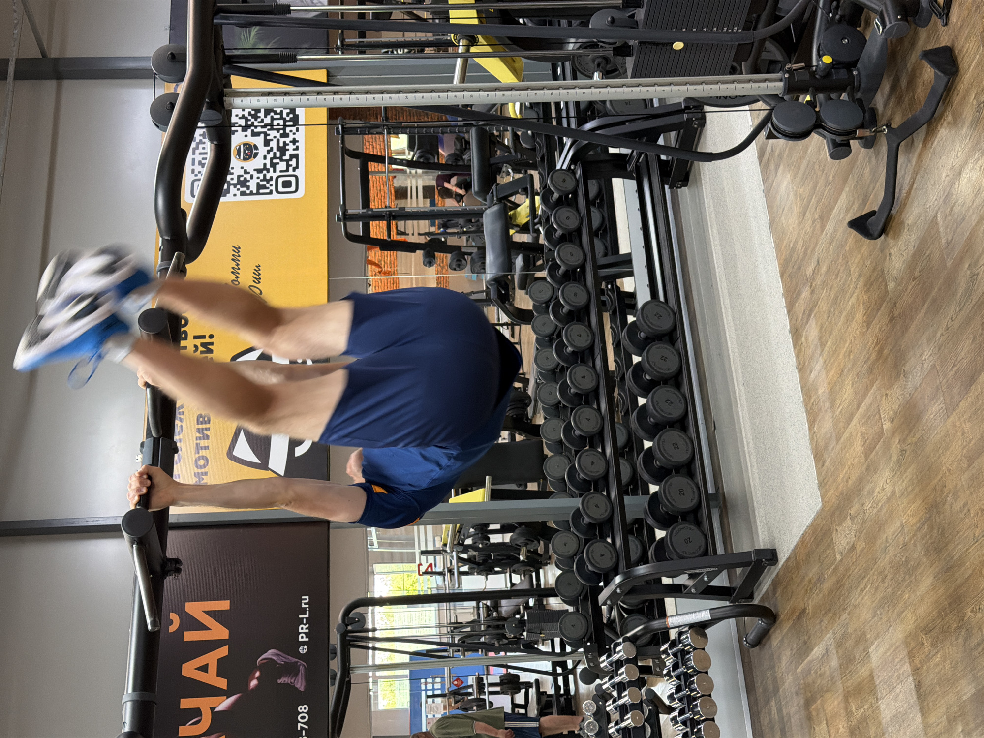 Here is a forensic description of the image:
**Scene:** An indoor fitness facility, likely in Chaikovsky, Russia.
**Subject:** An adult male is performing an inverted exercise on a pull-up bar. He is wearing blue shorts and white and blue sneakers.
**Objects:** Various gym equipment can be seen in the background, including dumbbell racks with a wide range of weight sizes, weight machines, pull-up bar and other fitness equipment. Signs are visible with Russian text, one with the word "ЧАЙ" (Chai) prominently displayed. There is a QR code, and the website "PR-L.ru" is mentioned.
**Actions:** The man is hanging upside down from the pull-up bar.
**Environment:** The gym appears to be well-equipped. A person is visible in the background. The floor is a wood-look laminate.