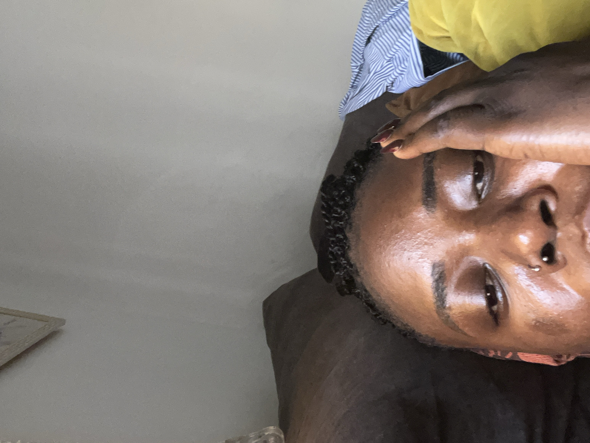 The image shows a close-up of a person's face and upper body, with their hand resting on their forehead. The person has dark skin and is wearing a nose ring. Their hair is styled in braids. The background is out of focus, but appears to be an indoor setting with a dark brown couch and some clothing, possibly a blue and white striped shirt and a yellow garment. The lighting suggests it might be daytime, possibly with natural light from a window. The image is cropped tightly on the person's face, with most of the surroundings obscured. There is no identifiable visual information within the image that would allow for the specific location of Abuja, Nigeria to be determined.