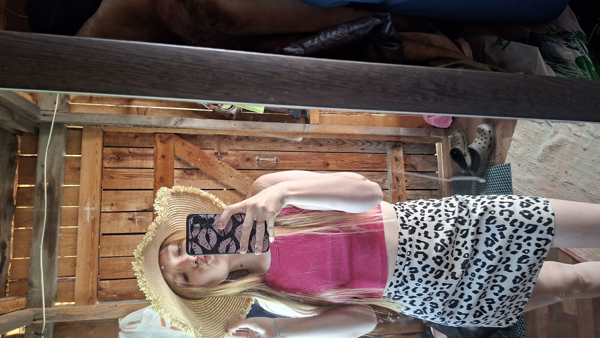 The image shows a reflection of a fair-skinned woman taking a selfie in a mirror, likely indoors. She's wearing a wide-brimmed straw hat, a pink crop top, and a white skirt with a black leopard print. She is holding a smartphone with a black case decorated with lips. The background visible in the reflection includes a wooden structure, possibly a shed or cabin interior. Above the reflection, the top of a shelf or surface with various items is visible. The location is presumably Большой Букор, Russia, based on the user's prompt.