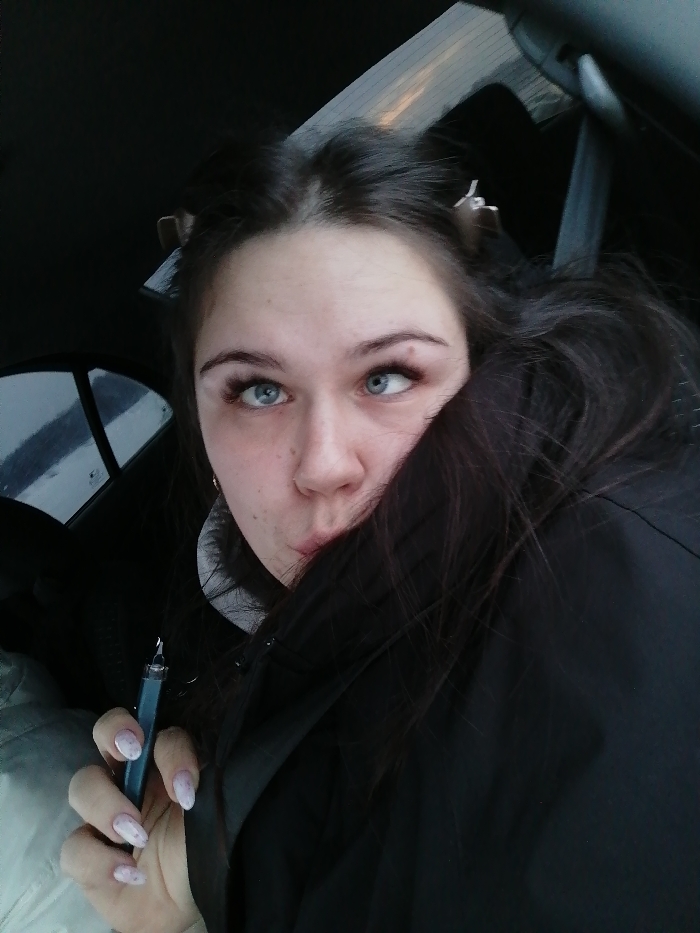 A young woman is in a car. She has dark hair with two clips in it. She's wearing a black coat pulled up around her neck, obscuring her face slightly. She holds a dark blue vape pen in her hand with light pink and white nails. She's looking to the side with a playful expression. The car window is visible in the background.