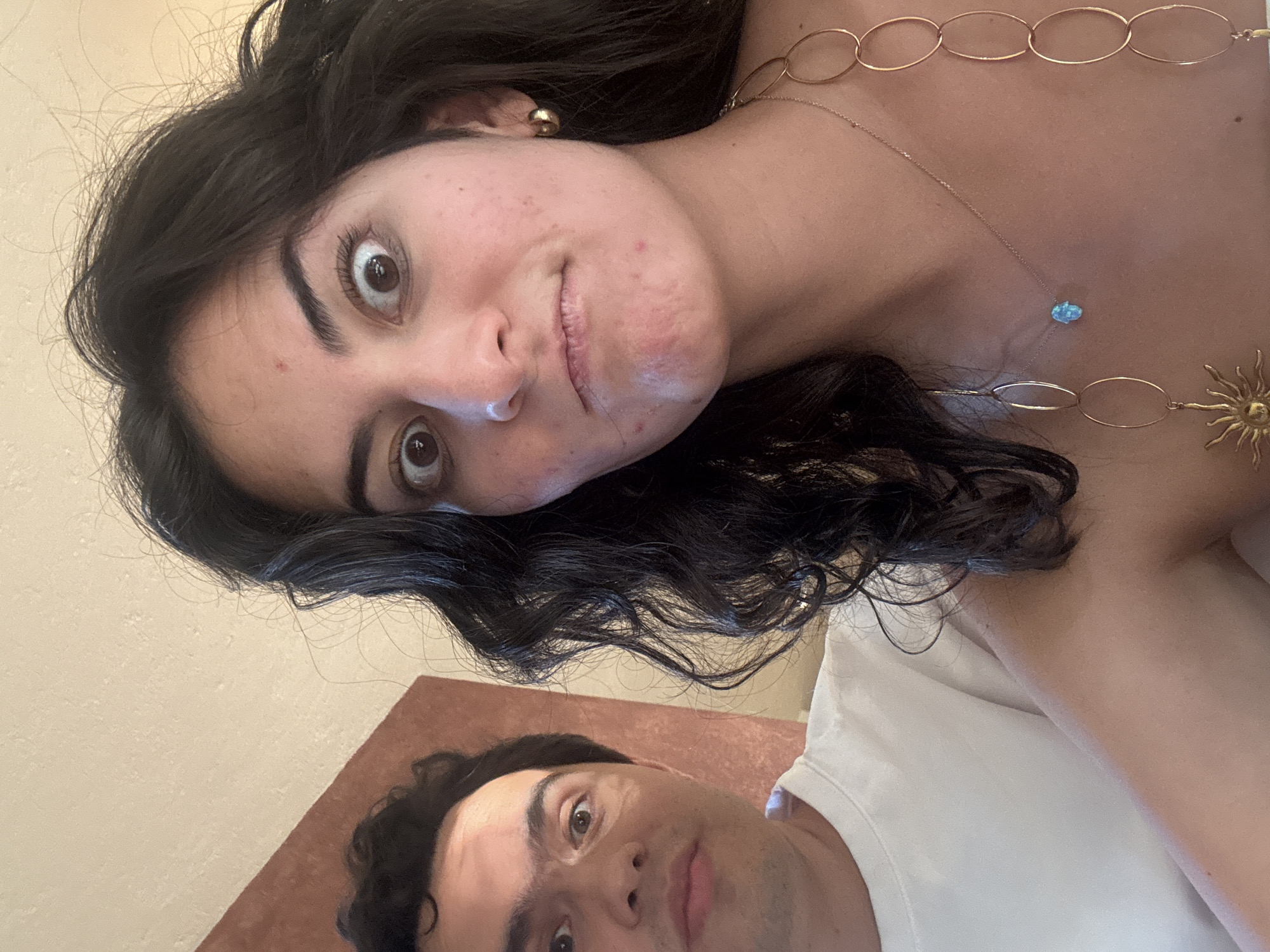 Two people are pictured in a selfie. The woman is positioned above and to the left of the man, tilting her head at an angle. She has dark, curly hair and a gold necklace. Her expression is playful, with wide eyes and a slight puckering of her lips. A gold body chain is visible on her upper chest and shoulder. The man is in the lower right corner of the image, wearing a white t-shirt. He looks towards the camera. The background appears to be an interior space with a beige-colored wall visible in the upper left. The location, San Bernardino Tlaxcalancingo, Mexico, is not discernible from the visual information in the image.