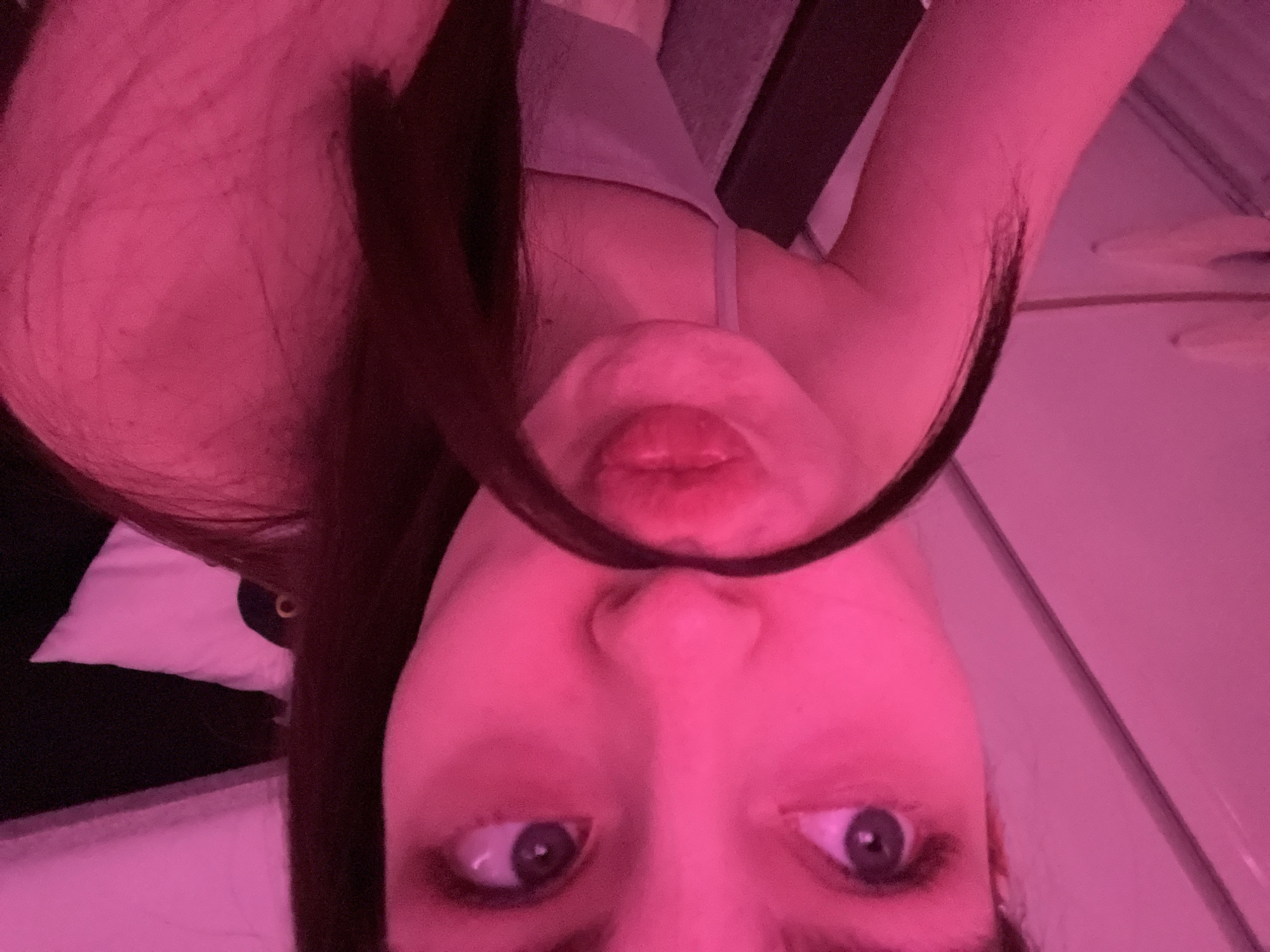 A selfie of a woman with fair skin, long brown hair, and blue eyes, taken with the camera rotated, showing a distorted reflection of her lips above her nose. The lighting is predominantly pink. A white pillow is visible in the background, along with indistinct furniture. The location cannot be determined from the given information.