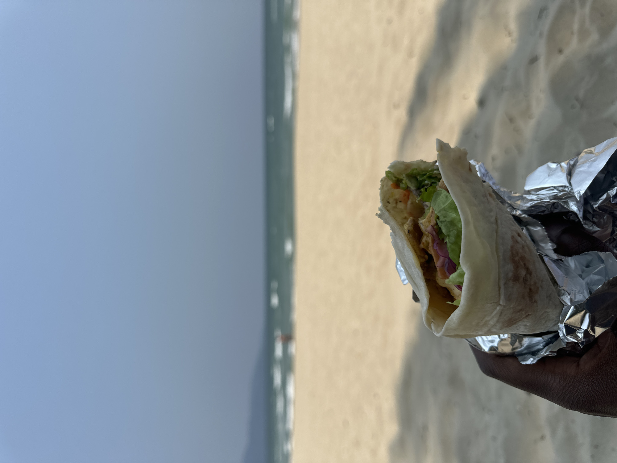 The image shows a partially consumed wrap, presumably a kebab or shawarma, held in a person's hand. The wrap contains lettuce, some unidentified ingredients, and is partially wrapped in silver foil. The background shows a beach and the sea, under a hazy sky. Based on the location information, the image was likely taken in Đà Nẵng, Vietnam. The wrap is the primary subject, held by a dark-skinned hand. The environment consists of a beach with sand and sea.